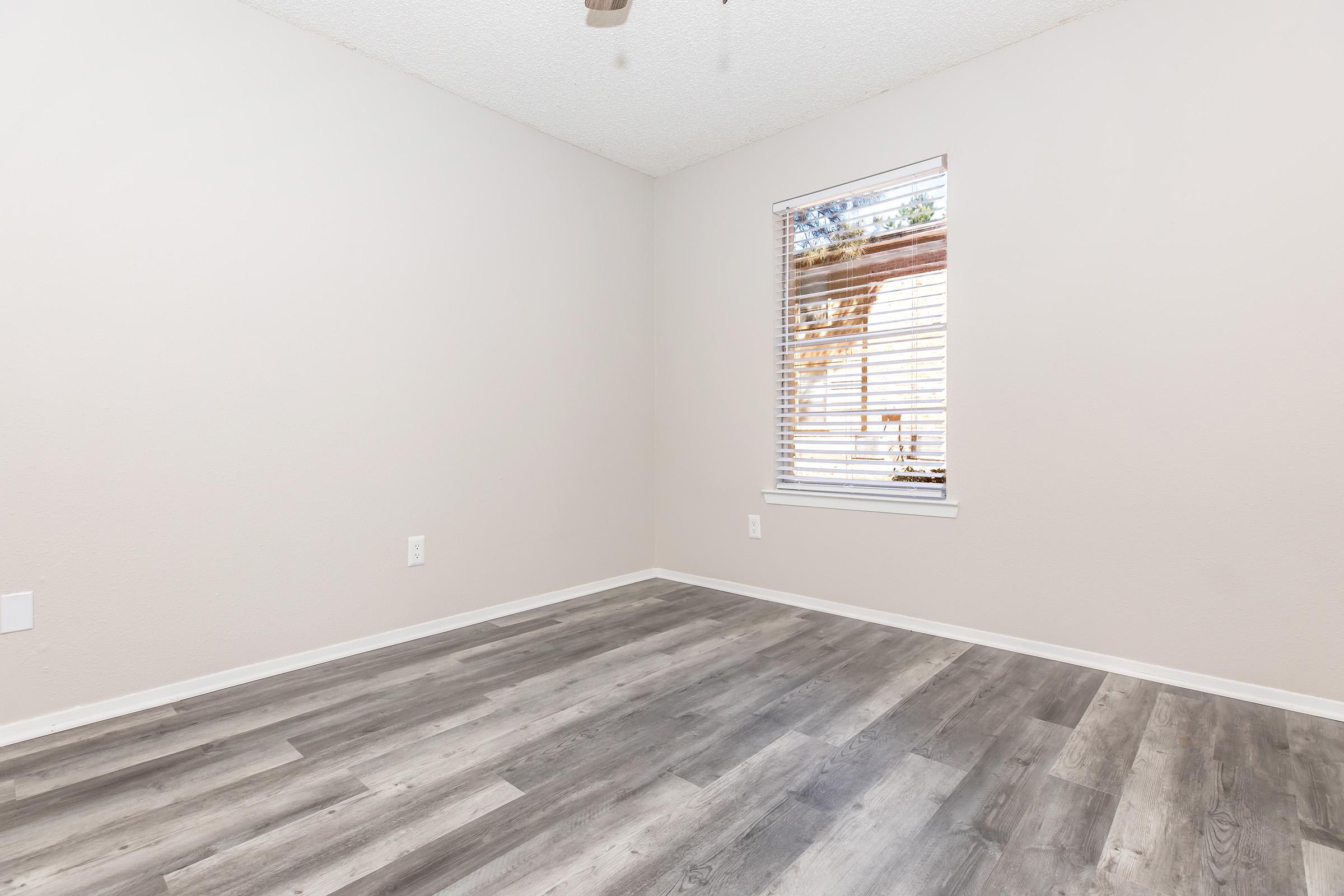 A vacant room with light gray walls, a ceiling fan, and white blinds on a window. The floor features a laminate design in shades of gray and brown. Natural light filters through the window, illuminating the space, which has no furniture or decorations.
