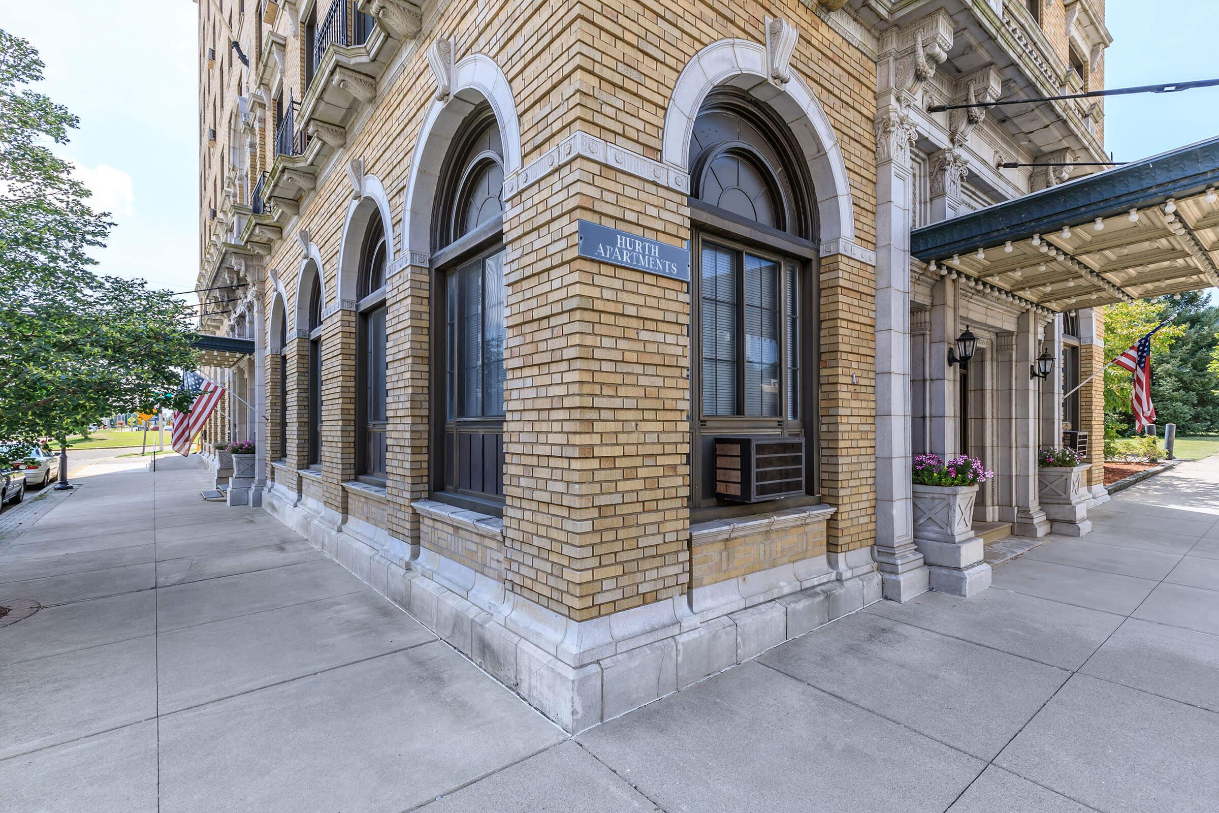 Corner view of a historic brick building featuring arched windows, decorative stone detailing, and flower planters. American flags are displayed along the façade, with a covered entrance visible. The surrounding area includes concrete walkways and greenery, suggesting a well-maintained exterior.