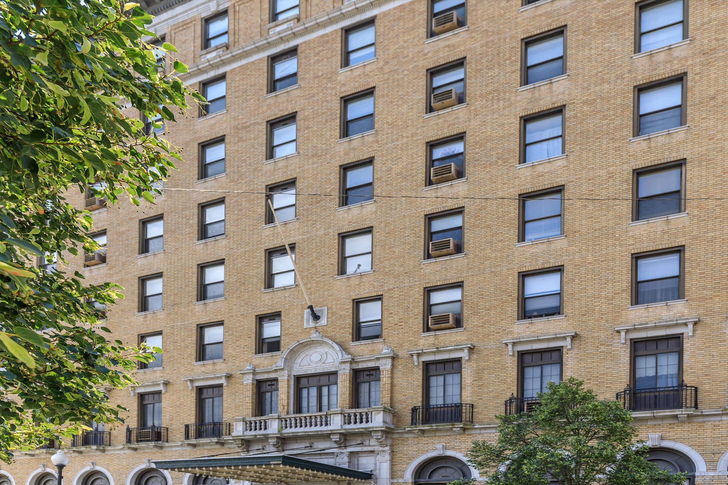 Brick building with multiple windows, featuring air conditioning units, topped with decorative architectural details. A canopy extends over the entrance, with greenery visible in the foreground. The facade shows a warm, golden hue typical of historic buildings.