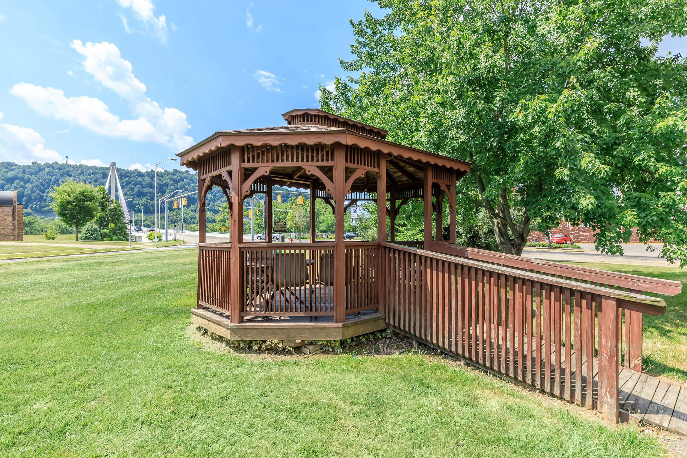A wooden gazebo with a slatted roof, surrounded by lush green grass and trees. The gazebo features a railing and an accessible ramp, set against a backdrop of blue skies and distant hills. The scene suggests a peaceful outdoor space suitable for relaxation or gatherings.