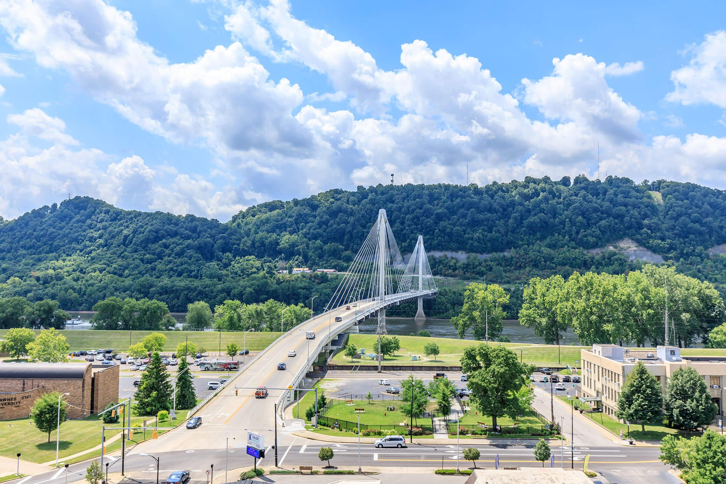 A scenic view of a cable-stayed bridge spanning a river, surrounded by lush green hills. The sky is partly cloudy with blue patches. The foreground features a roadway with parked cars, trees, and buildings, creating a lively urban atmosphere near the bridge.