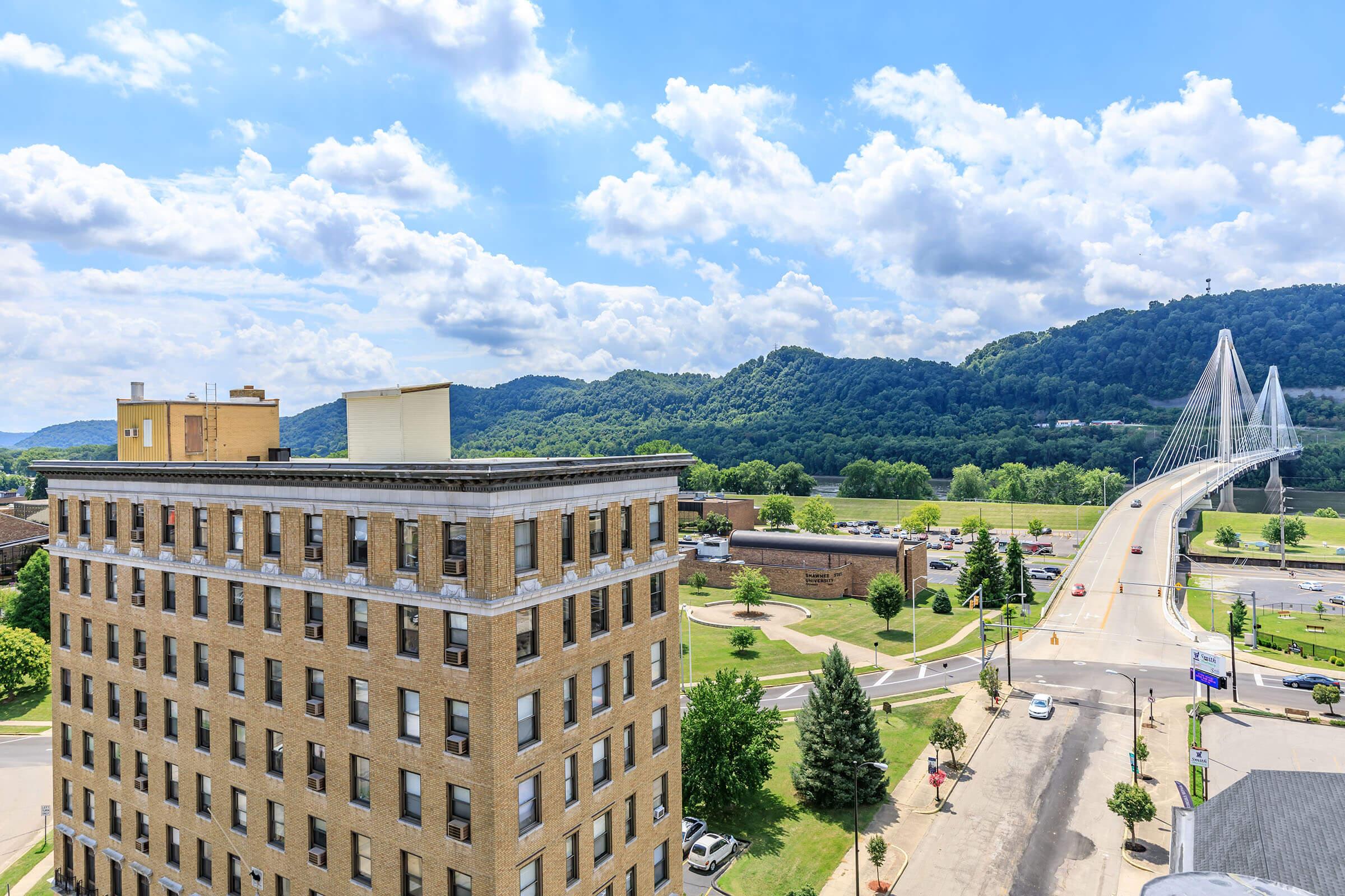 View of a cityscape featuring a multi-story brick building in the foreground, with a modern cable-stayed bridge crossing a river in the background. Lush green hills rise on either side of the river under a blue sky dotted with fluffy white clouds. The scene conveys a blend of urban and natural landscapes.