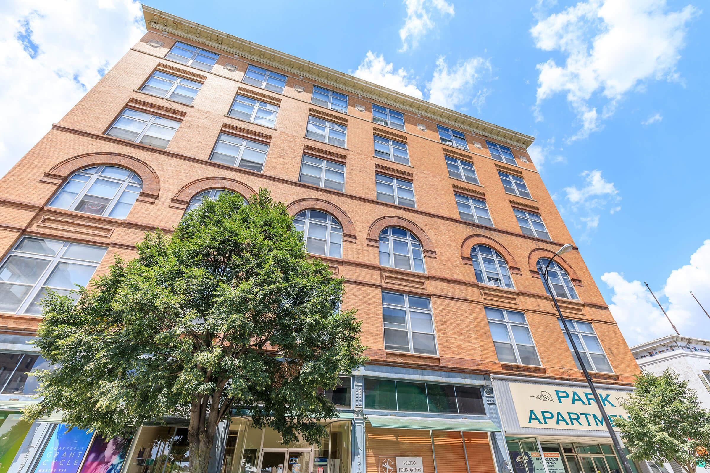 A five-story brick apartment building with large arched windows and a green tree in the foreground. The building has a sign that reads "Park Apartments," and the sky is bright with fluffy clouds.