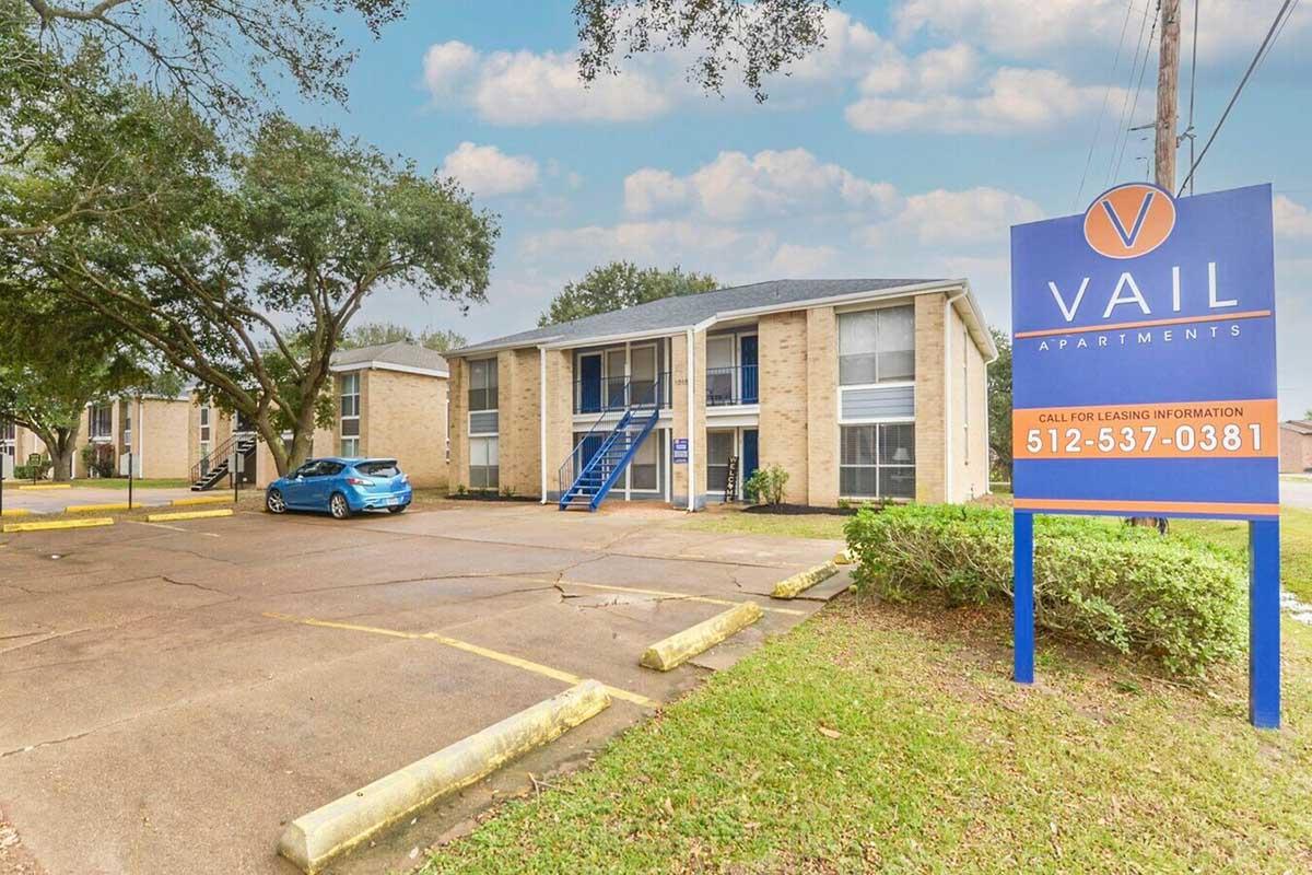 A view of Vail Apartments featuring two-story buildings with a blue car parked in the foreground. The entrance is marked by a prominent sign displaying the name and contact information for leasing inquiries. The surroundings include green grass and trees under a cloudy sky.