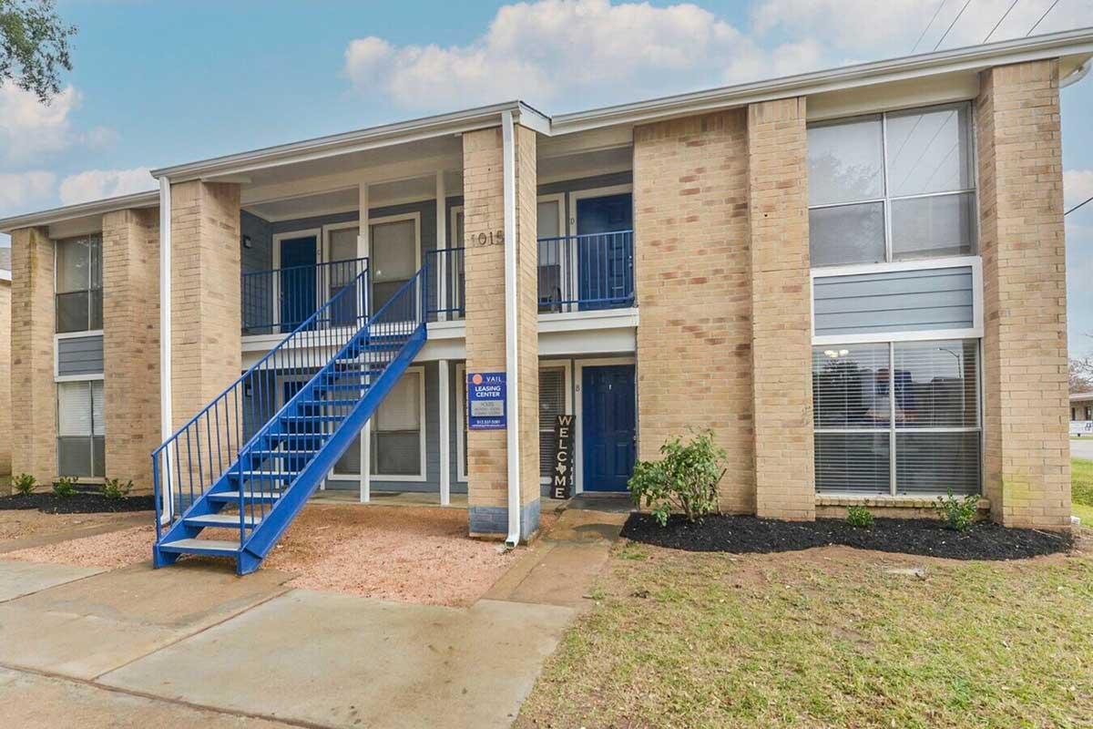 Exterior view of a two-story brick apartment building, featuring blue railings and stairs. The entrance has a welcome sign, and the surrounding area is landscaped with small plants, gravel, and grass. Large windows allow for natural light, creating an inviting atmosphere.