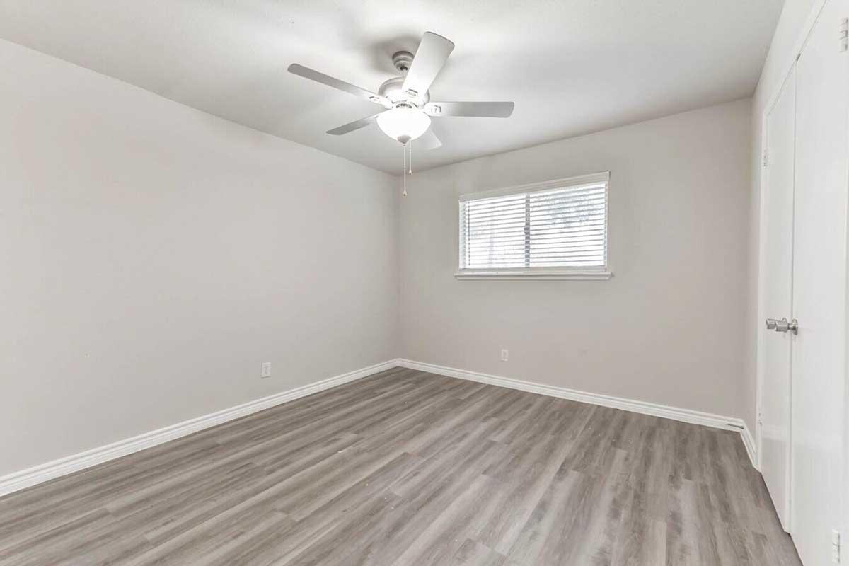 A bright, empty room with light gray walls and a ceiling fan. It features a window with blinds, allowing natural light, and has light-colored laminate flooring. The room has a simple, clean aesthetic suitable for various purposes, with a door on one wall. No furniture is present.