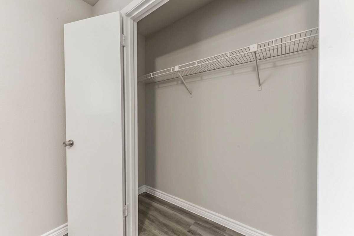 An empty closet with a white door slightly open, showcasing a clean interior with light-colored walls and a shelf for storage. The floor features a wood-like design, emphasizing the spacious and minimalist appearance of the space.
