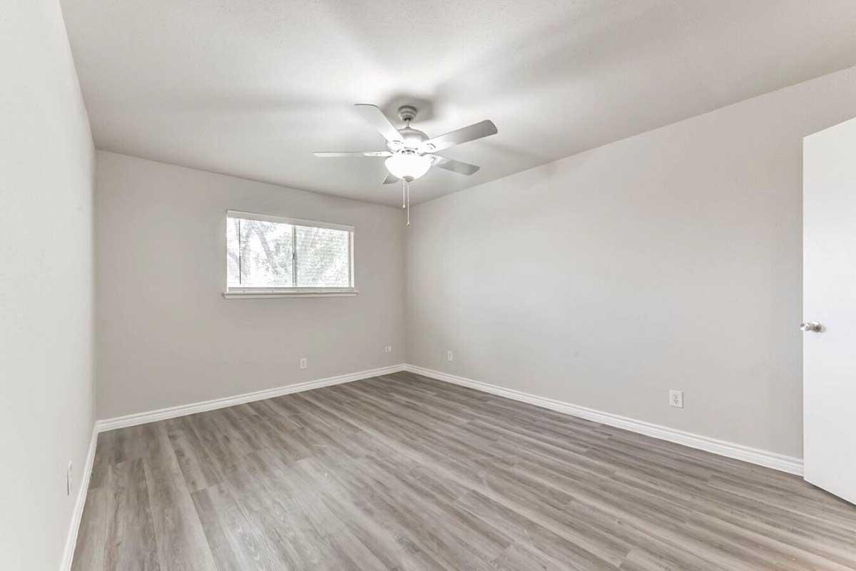 A minimalistic bedroom with light grey walls and wood-like flooring. It features a ceiling fan, a window with natural light, and one door. The room is empty, providing a spacious feel.