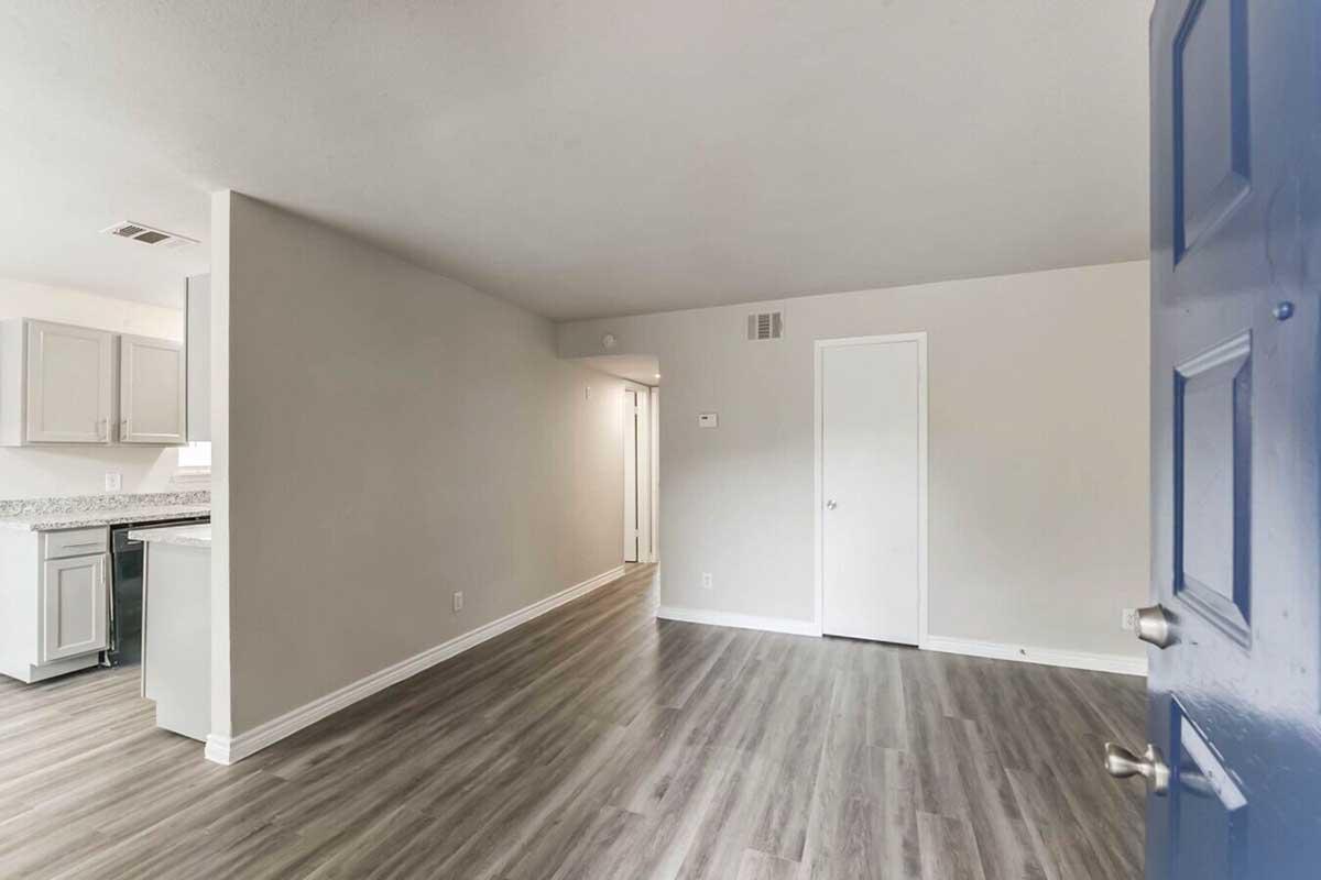 A view of an interior space from a doorway, featuring light-colored walls and a wooden floor. To the left, there is a kitchen area with light cabinetry, and opposite, a closed door leading to another room. The space appears bright and modern, with natural light enhancing the open layout.