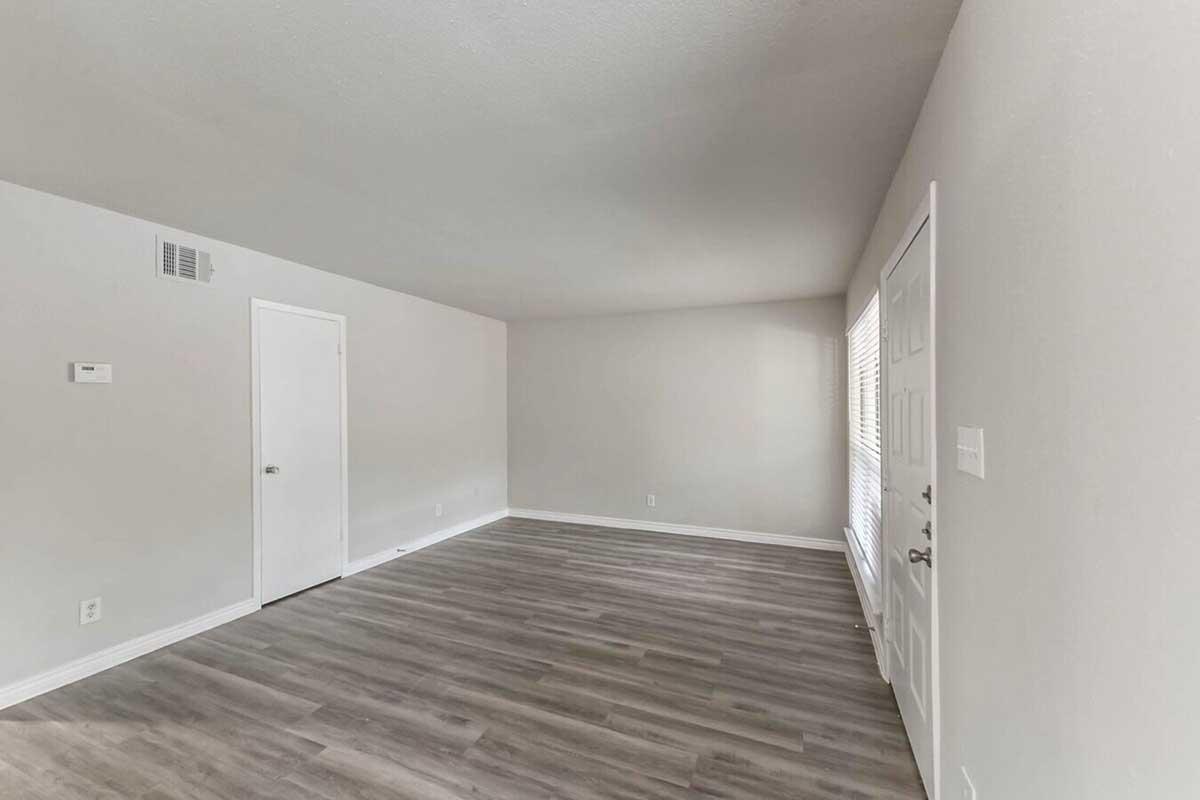 A spacious, empty room with light gray walls and a smooth wooden floor. A single door on the left leads to another area, while a white door on the right appears to be a closet. Natural light streams in from a window on the far wall, illuminating the uncluttered space.