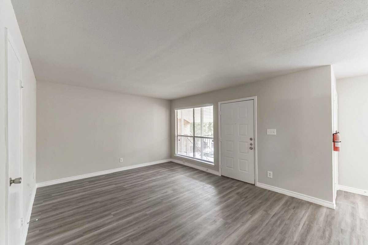 A bright, empty living room with light gray walls and laminate flooring. There's a large window with blinds on one wall, providing natural light, and a white door leading outside. The room has minimal decor and a fire extinguisher mounted near the entrance.