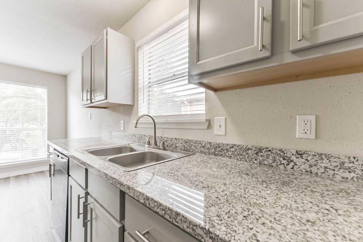Modern kitchen featuring gray cabinets, a granite countertop with a double sink, and a window allowing natural light. The space has a clean, minimalist design with neutral colors and a wooden floor.