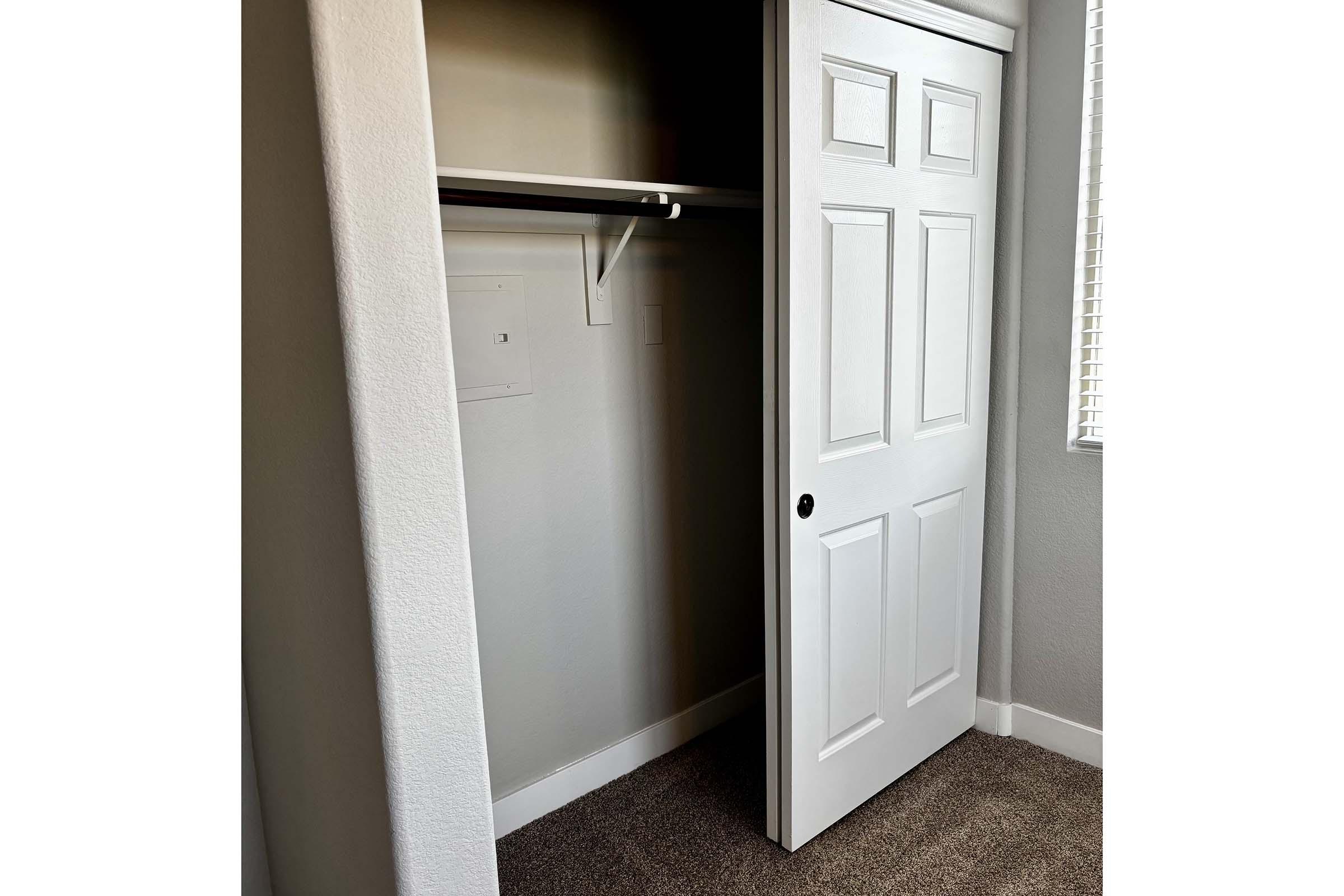 A partially open closet door reveals an empty closet with a shelf and light gray walls. The floor is covered in beige carpet, and there is a window with blinds casting soft light into the space. The overall atmosphere is neat and minimalistic.