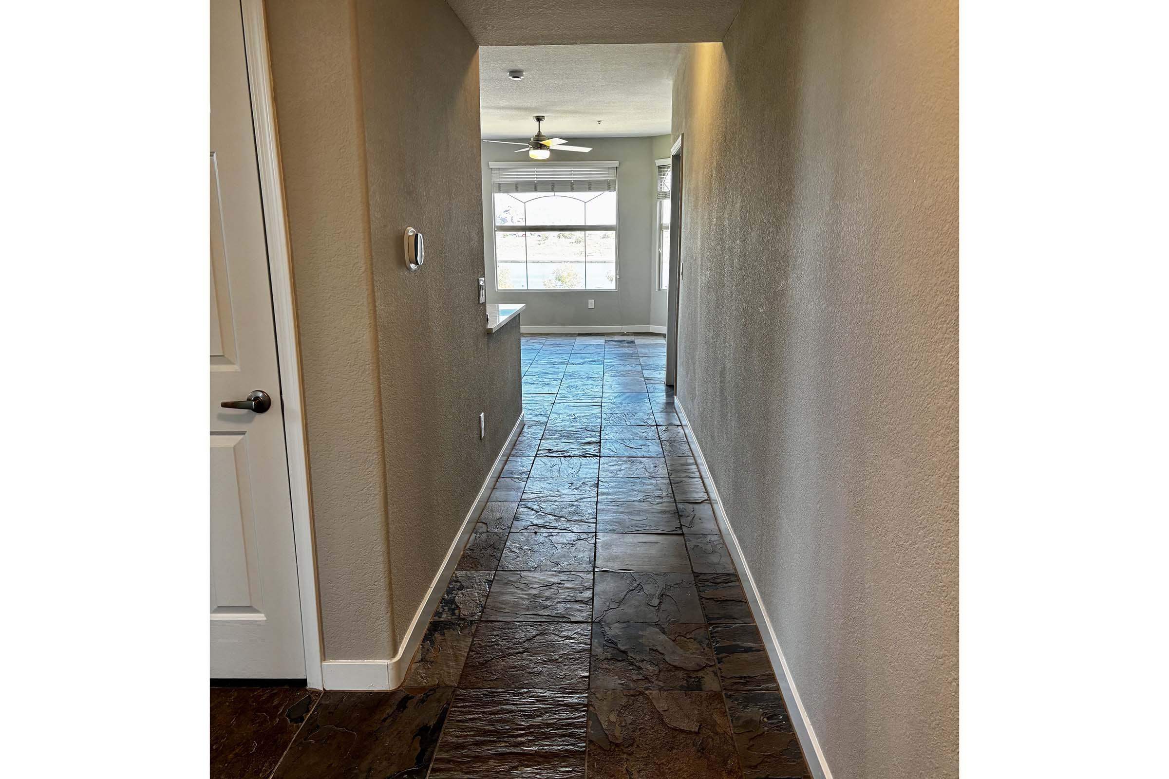 A long hallway with textured stone flooring, leading towards a brightly lit room with a large window. The walls are painted in a light neutral color, and a ceiling fan is visible in the distance. The entrance to a room is on the left side.