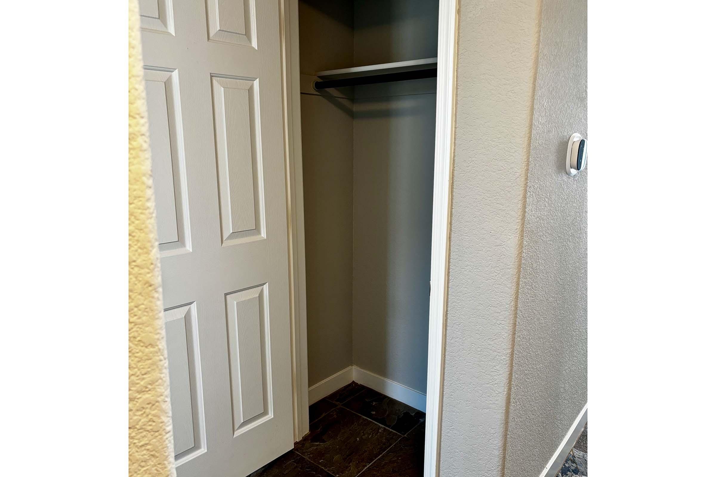 A partially open closet door revealing an empty closet space with a shelf. The walls are painted light gray, and the floor features dark tiles. A light switch is visible on the right wall.