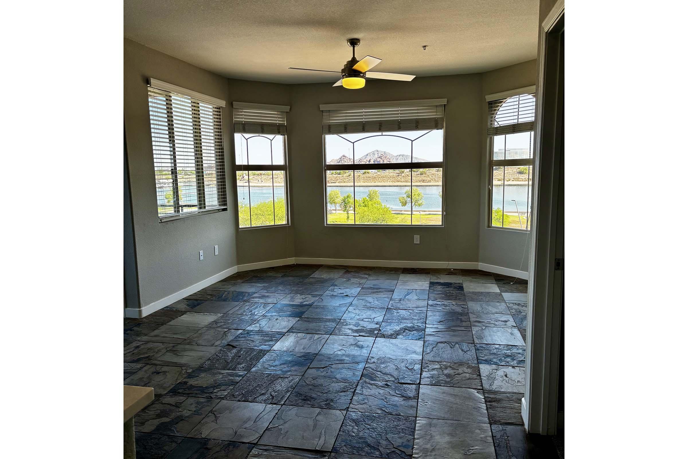 A spacious room with large windows offering a view of a body of water and greenery. The floor is covered with elegant slate tiles. The room features a ceiling fan and is illuminated by natural light, creating a bright and airy atmosphere.