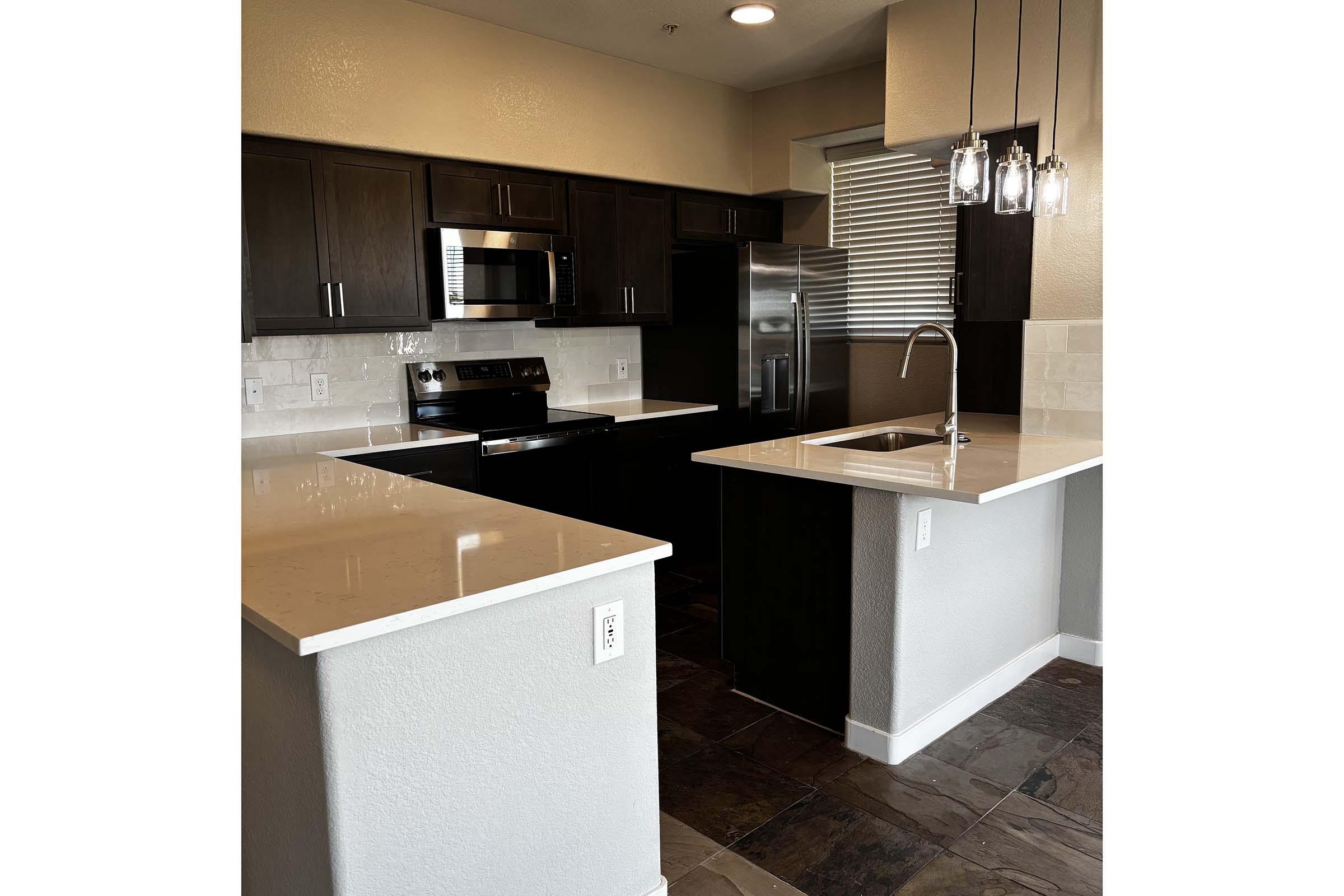 A modern kitchen featuring sleek dark cabinetry, stainless steel appliances, and a spacious island with a white countertop. The area is well-lit with pendant lighting and has a neutral color palette, complemented by a tile floor. Large windows provide natural light in the space.