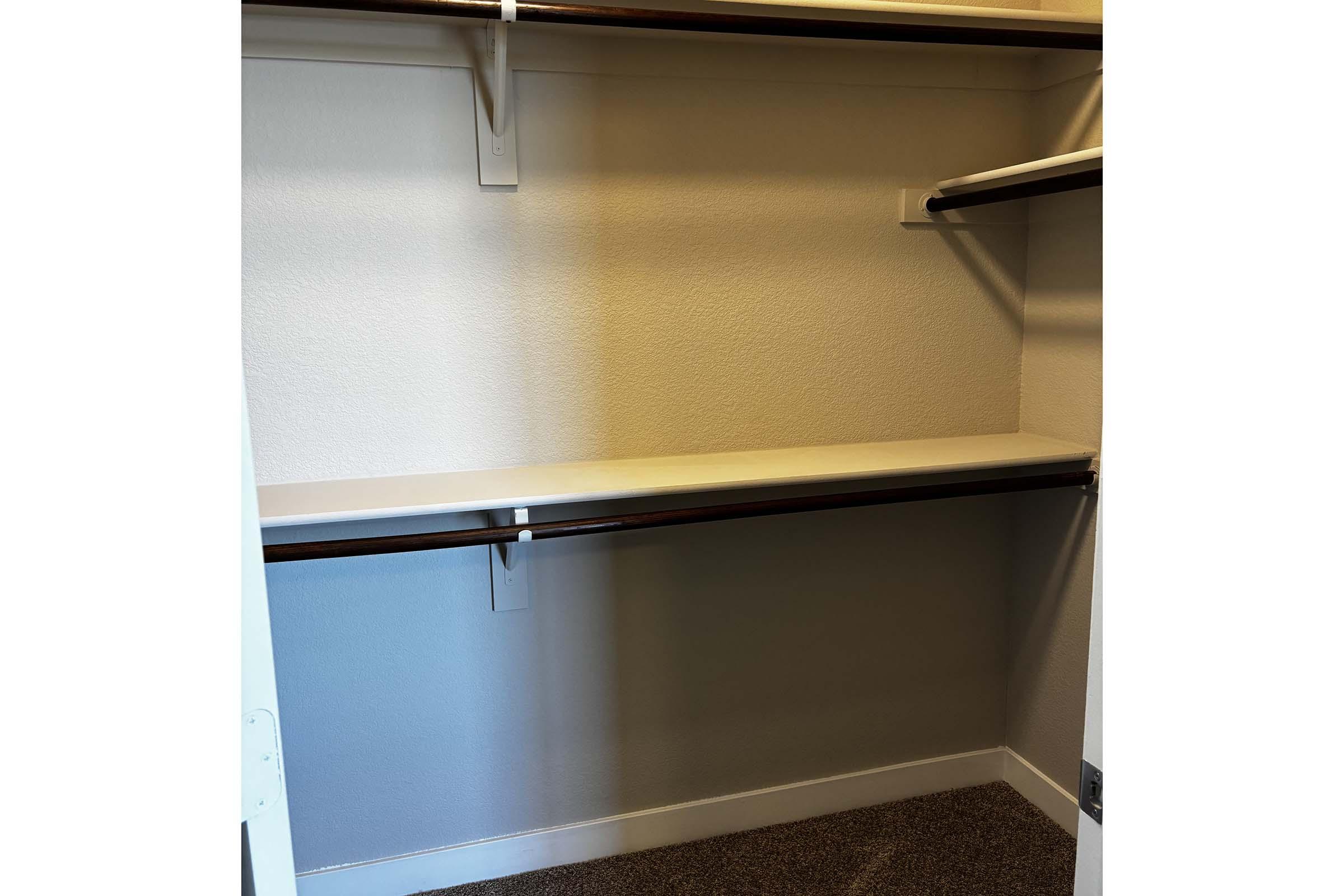 A vacant closet with two shelves. The upper shelf is made of wood and mounted on the wall, while the lower shelf is a simple surface without any items. The walls are painted light, and the carpet is a neutral color, creating a clean and organized space.