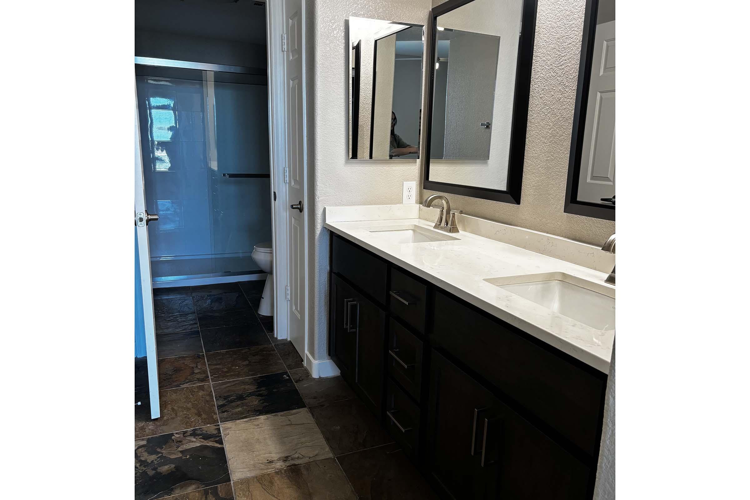 A modern bathroom featuring a double vanity with two sinks, dark wood cabinets, and two mirrors above the sinks. The floor is tiled with a mix of dark and light colors. In the background, a glass shower enclosure is visible, along with a partially closed bathroom door.