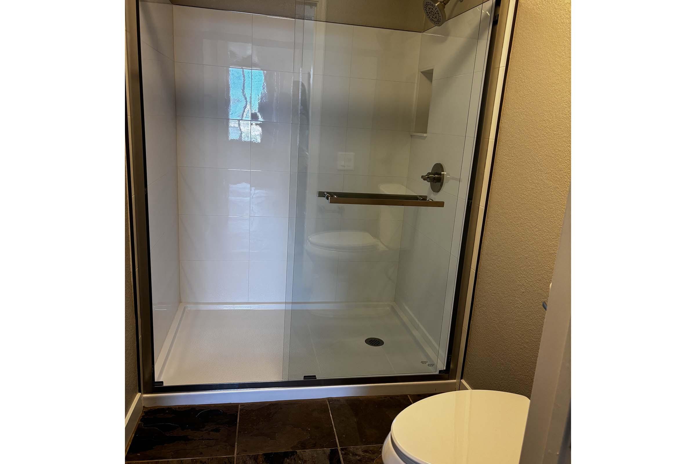 A modern bathroom featuring a glass shower enclosure with white tiled walls, a built-in shelf, and a small circular drain on the floor. A white toilet is positioned nearby on dark tiled flooring, and natural light is filtering through a window.