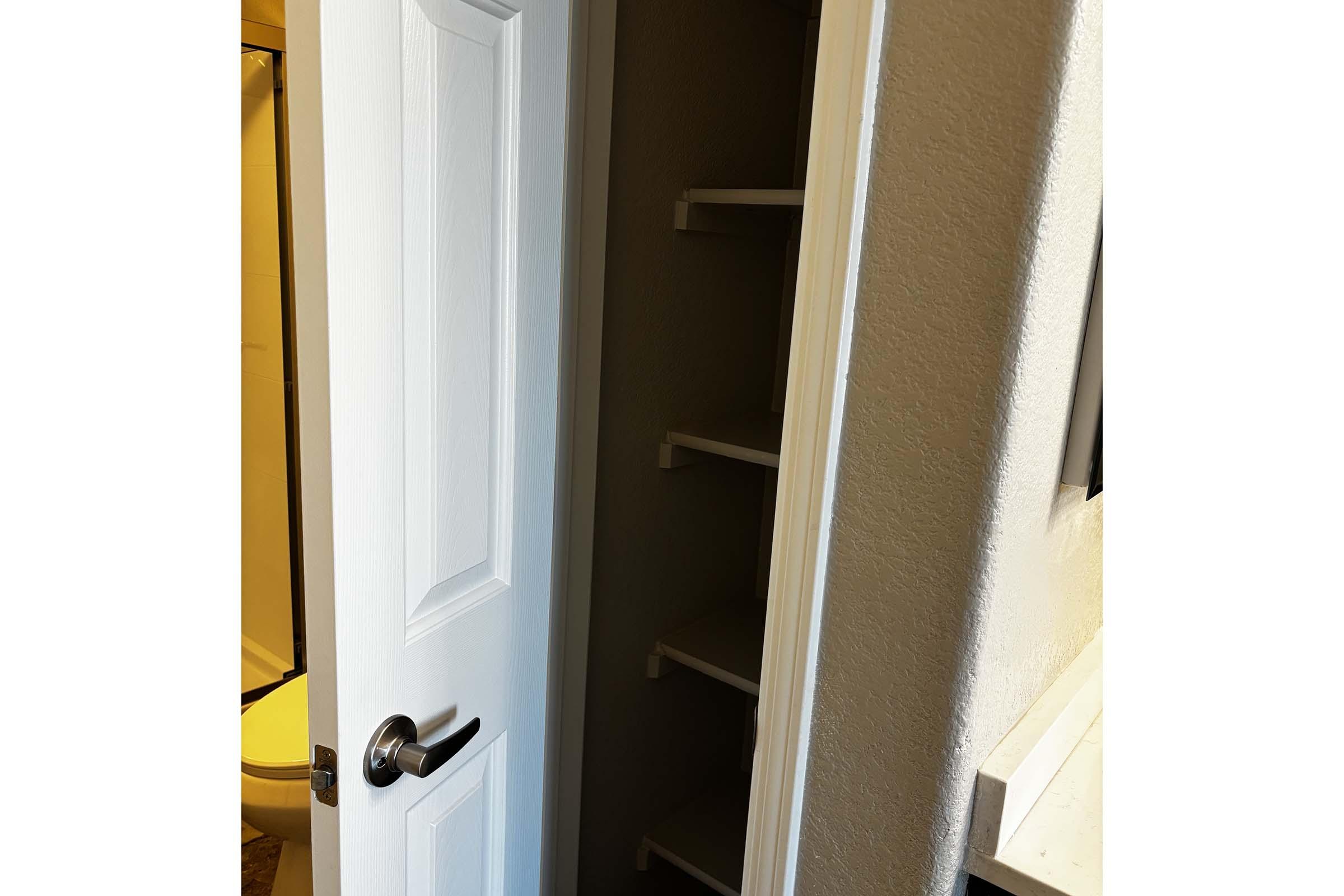 A partially open white door leads into a small closet or storage area with empty shelves against a light gray wall, revealing a glimpse of a bathroom with yellow tiles in the background. The floor is not visible, and the overall atmosphere appears tidy and organized.