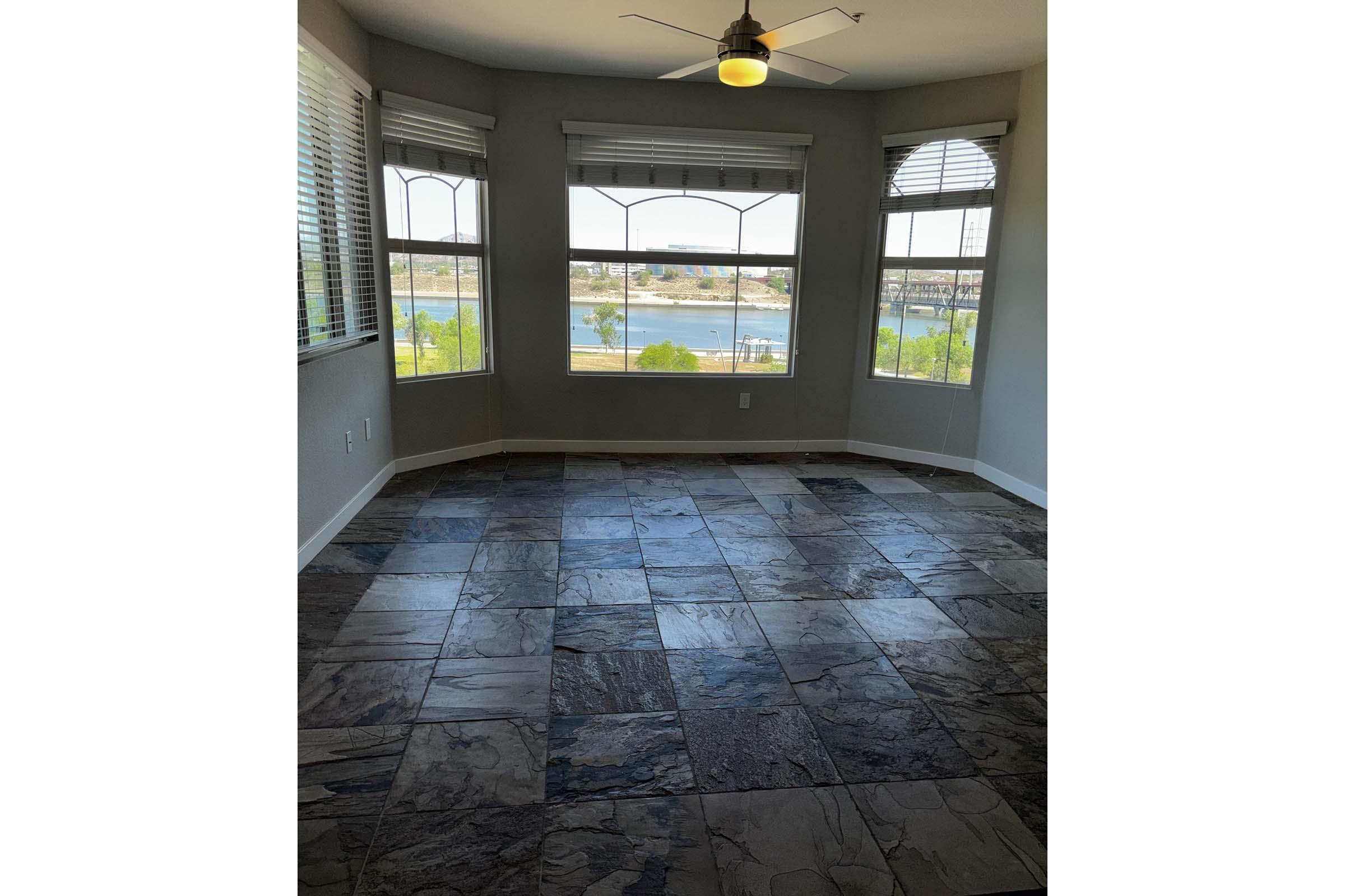 Interior view of a room with large windows showcasing a waterfront view. The floor is made of dark, patterned tiles, and there is a ceiling fan overhead. Natural light fills the space, highlighting the clean lines and contemporary design.