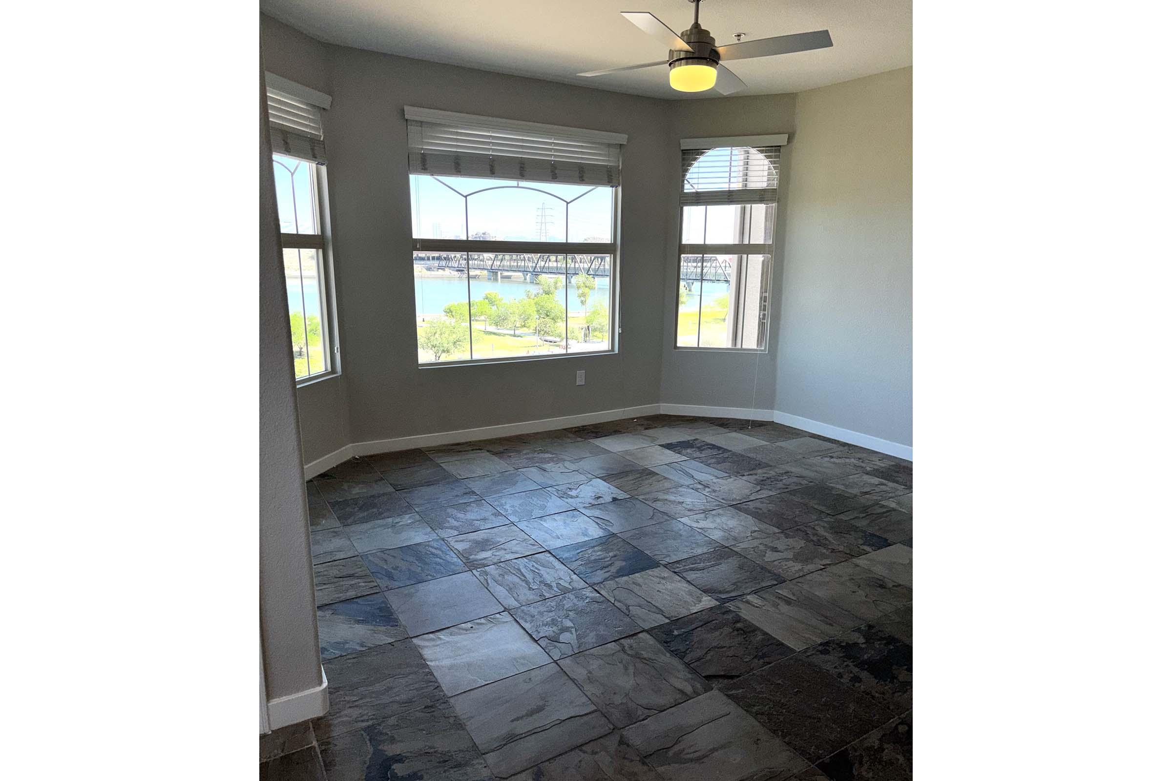 A well-lit room with large windows offering a view of a river or lake. The floor is covered in dark, glossy tiles, and there is a ceiling fan. The walls are painted in a light color, creating a spacious and airy feel. The overall ambiance is modern and inviting.
