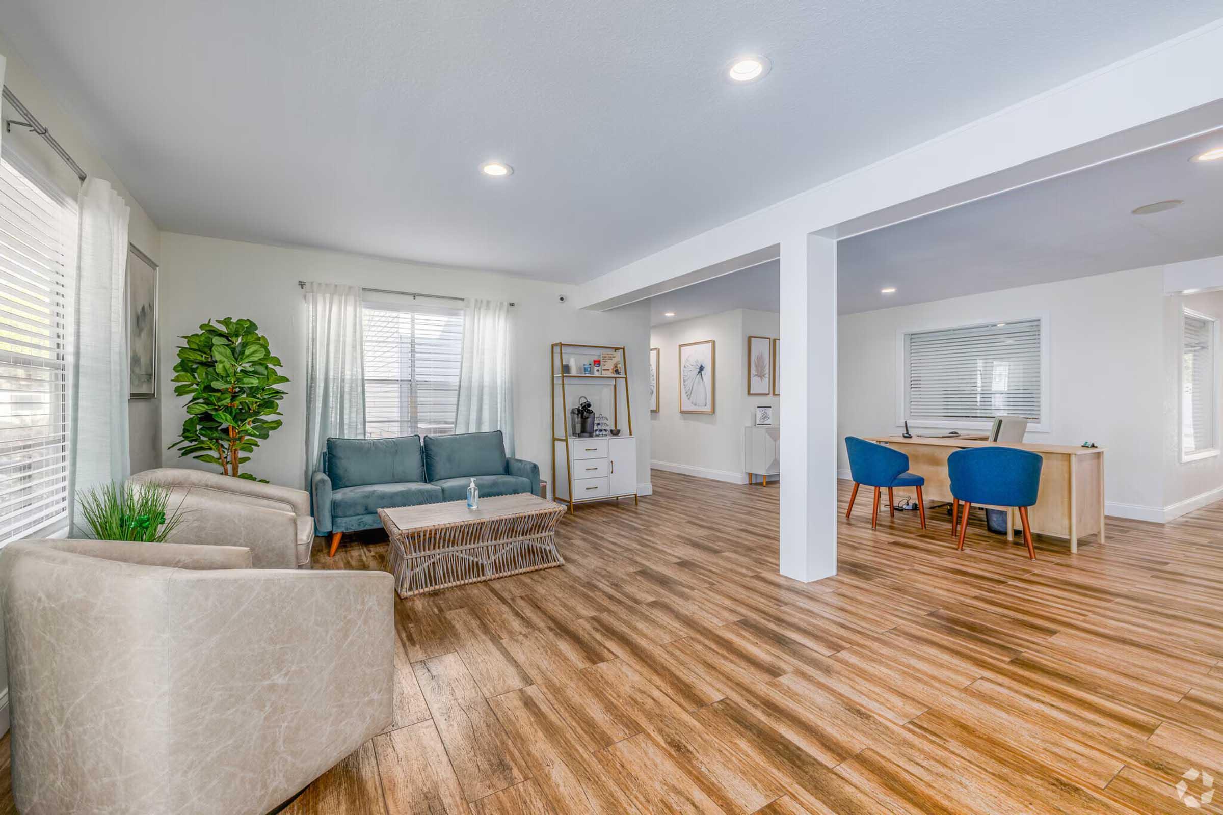 Spacious and modern living area featuring a light blue sofa and two beige armchairs. A coffee table sits in the center, and large windows allow natural light to filter in. There’s a small dining area with blue chairs and a wooden table in the background, accented by decorative plants and wall art.