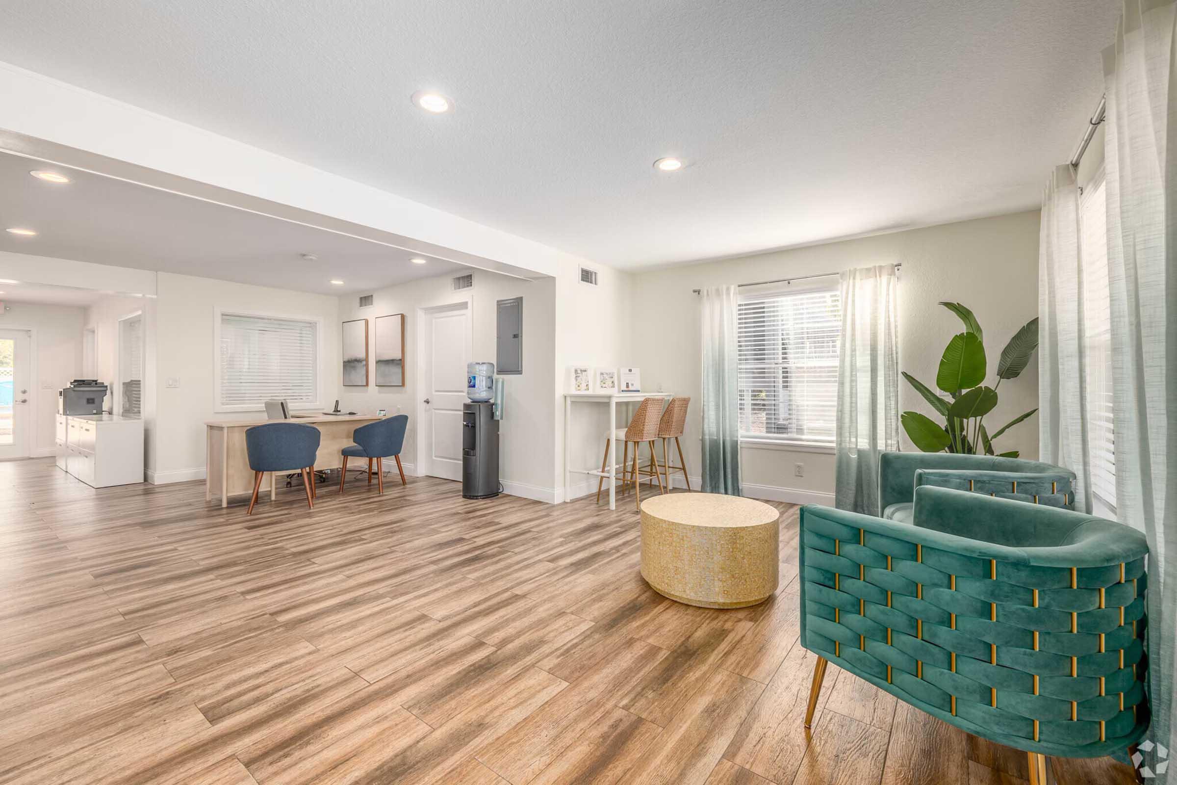 A modern, open-concept living space featuring light wood flooring, bright walls, and large windows. The room includes a cozy green armchair set, a round coffee table, a small dining area with blue chairs, and a kitchen view in the background, creating a welcoming atmosphere.