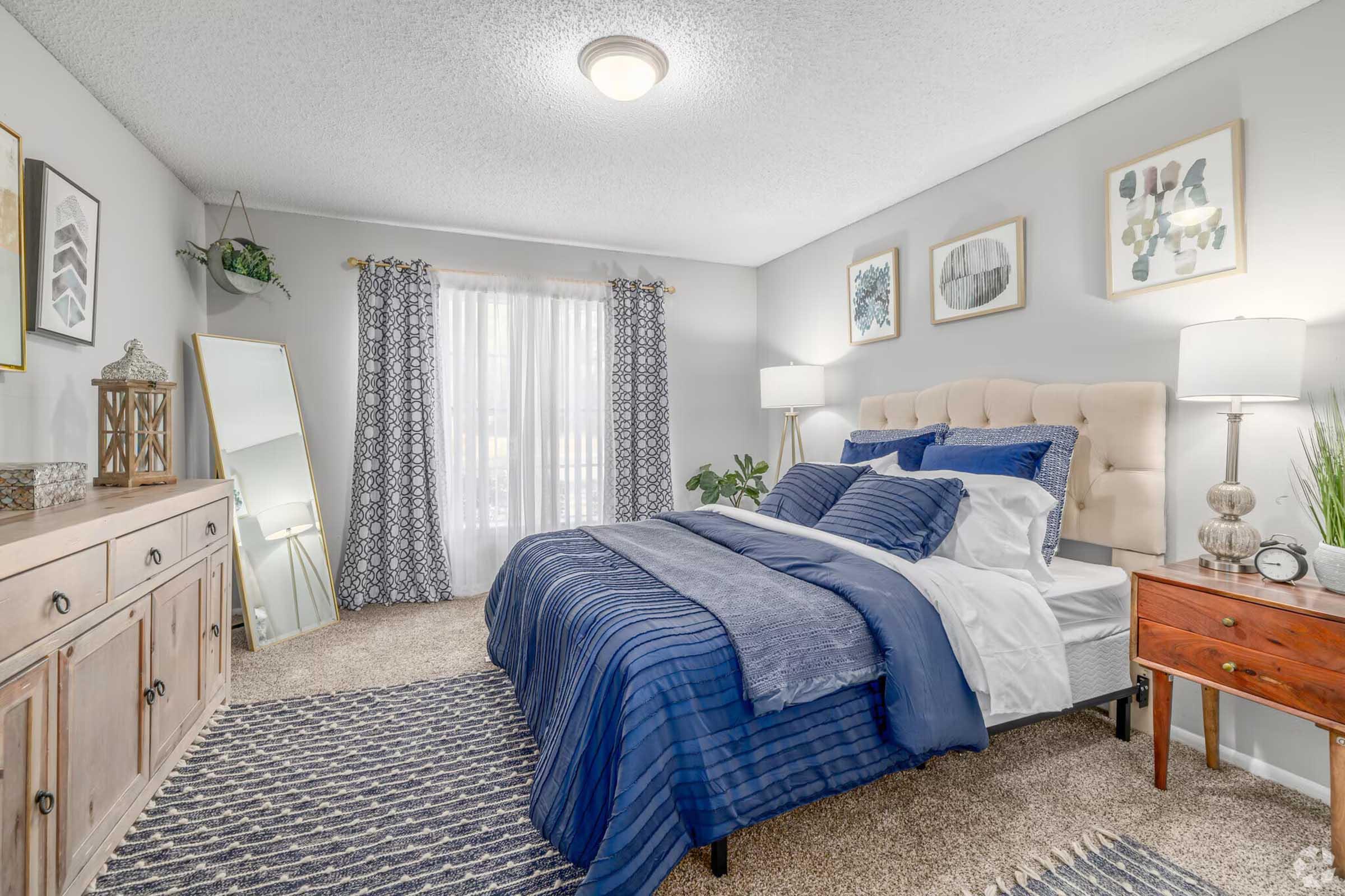 A cozy bedroom featuring a king-sized bed with navy blue bedding, a light-colored tufted headboard, and decorative pillows. The room includes a wooden dresser, a bedside table with a lamp, a large mirror, and patterned curtains. Natural light filters through a window, creating a welcoming atmosphere.