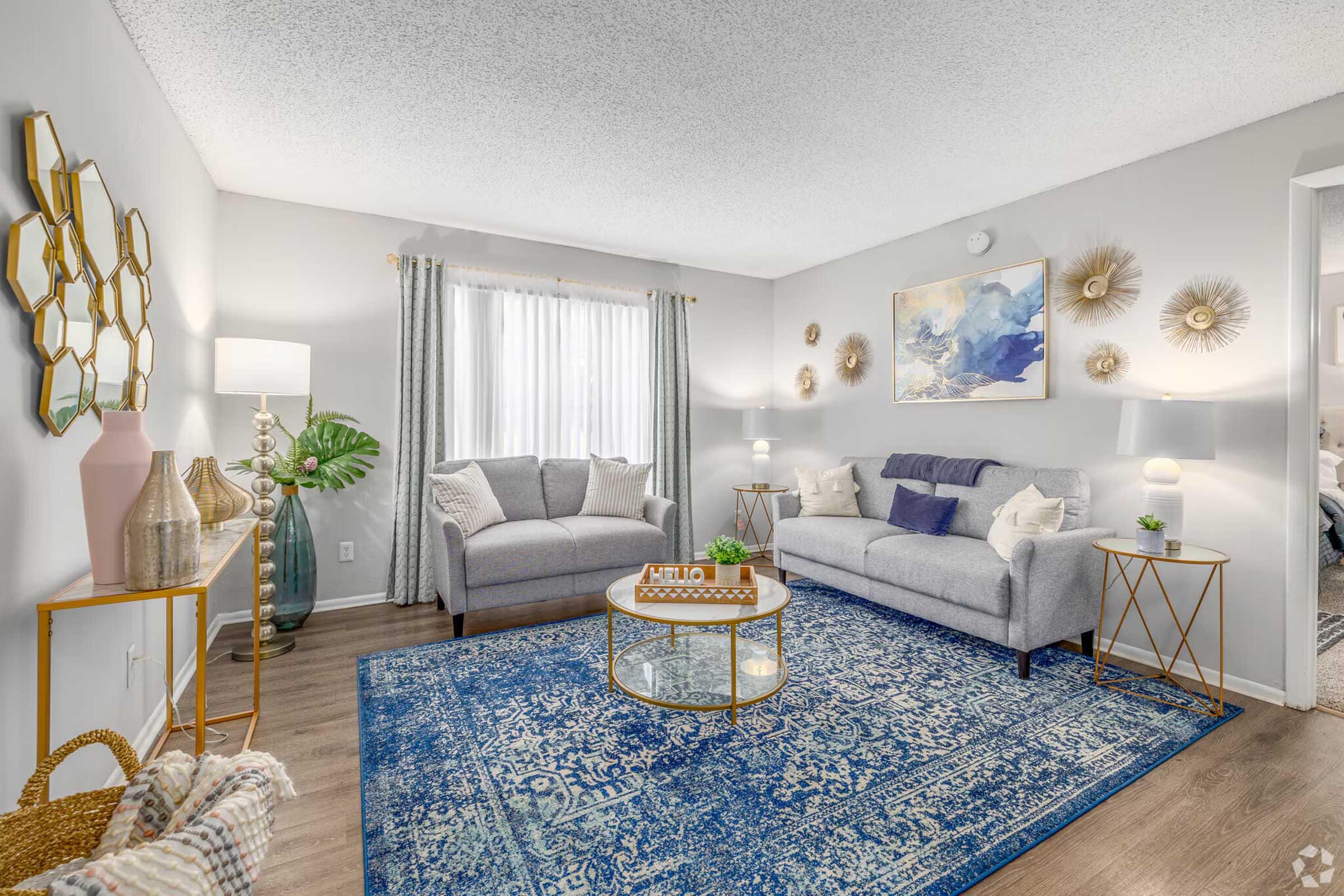 A cozy living room featuring a light gray sofa and loveseat, accented with decorative pillows. There is a large blue area rug on the floor, a coffee table with decorative items, and a plant. The room has bright curtains, wall art, and various wall decorations, creating a warm and inviting atmosphere.