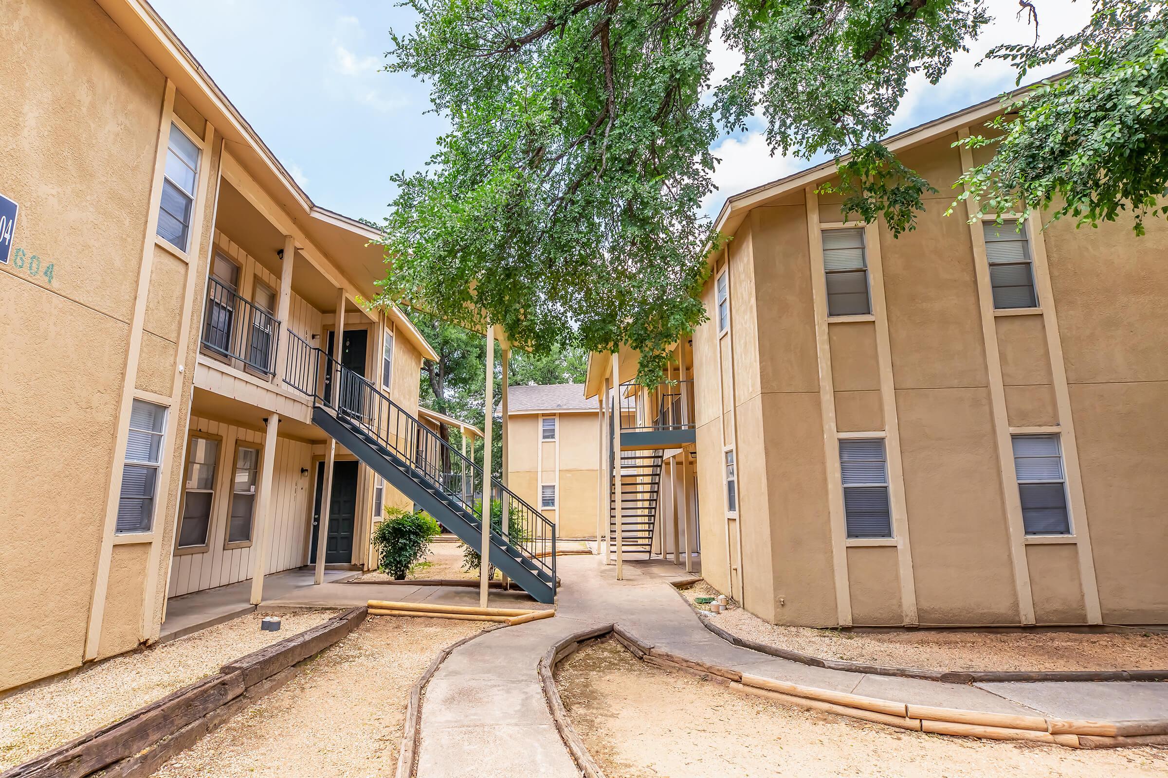 A view of a mid-rise apartment complex with two buildings surrounding a landscaped courtyard. The buildings feature multiple balconies and staircases, while a winding path connects the units. Trees provide shade, and there is a gravel pathway leading through the area.