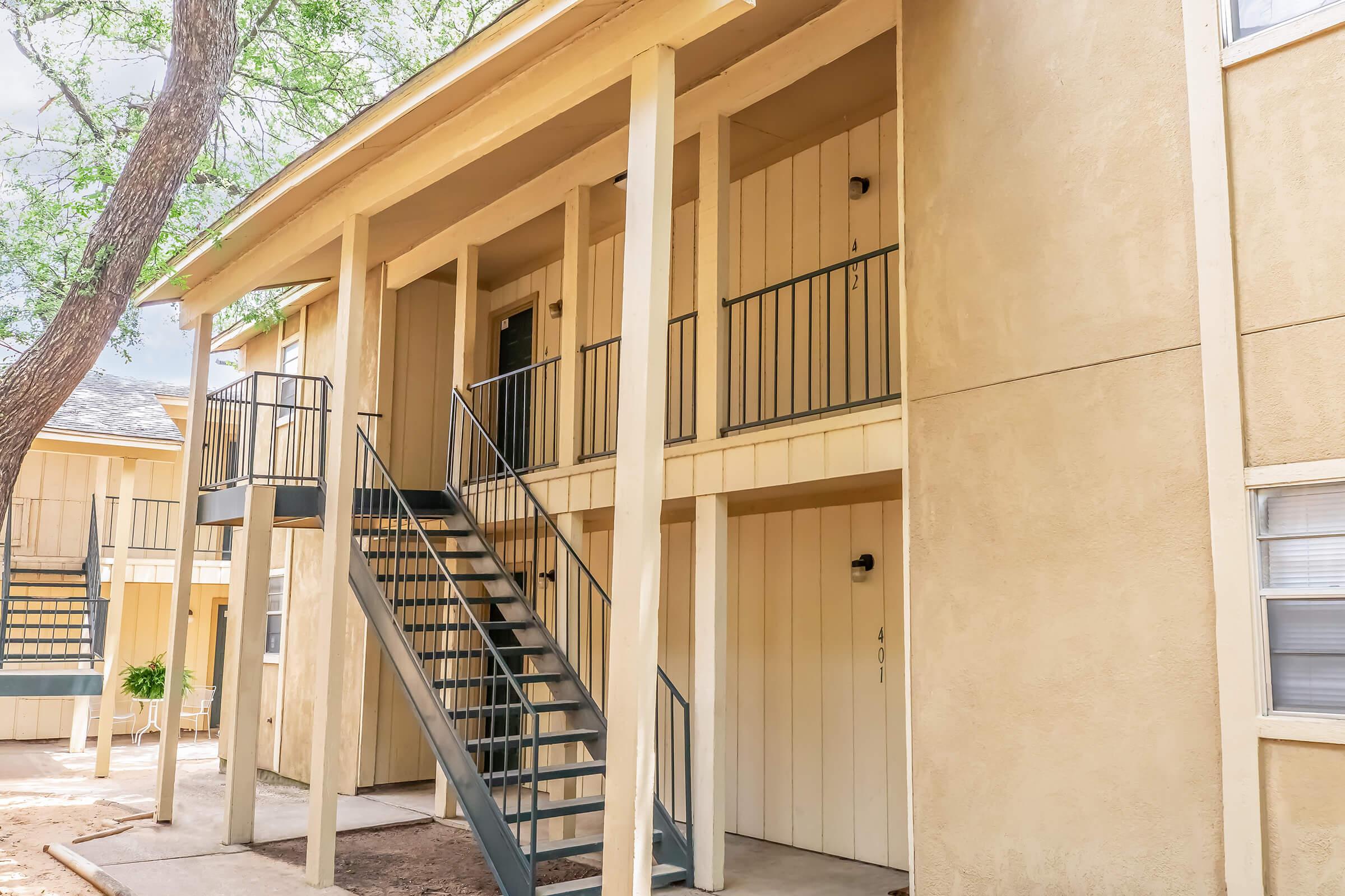 Two-story beige apartment building with a staircase leading to upper units. The building features wooden siding, metal railings, and numbered doors. A tree is visible nearby, providing shade to the area.