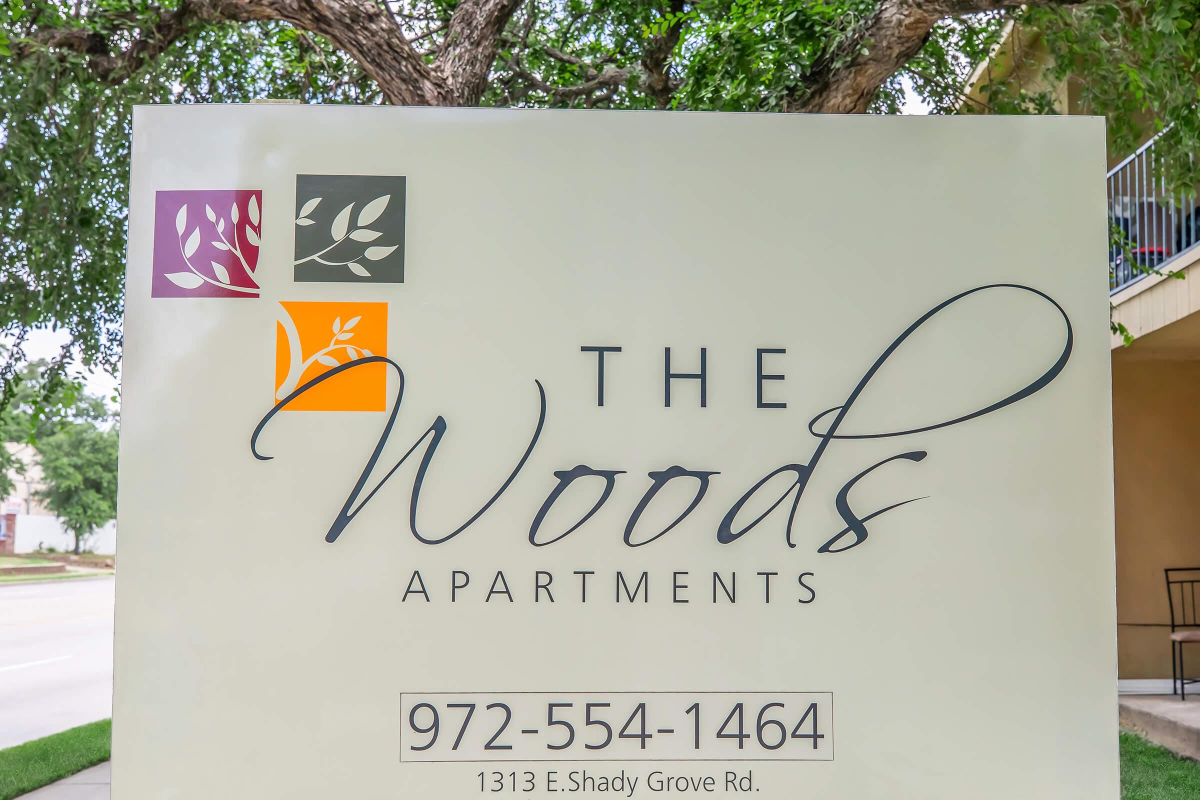 Sign for The Woods Apartments, featuring the name prominently in elegant script. The sign includes colorful leaf graphics in the top corners, and contact information along with the address: 1313 E. Shady Grove Rd. The overall design is modern and inviting.