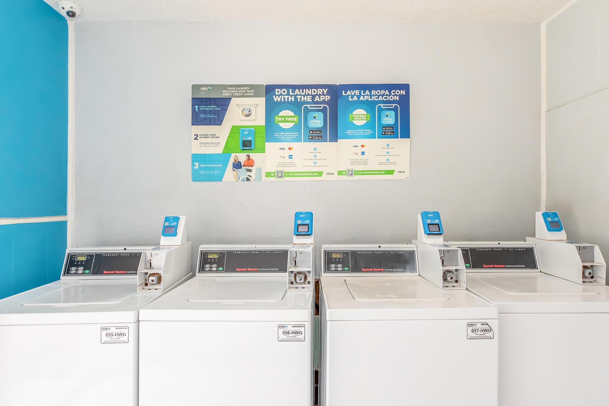 A clean laundromat featuring several white washing machines with digital displays. A wall-mounted sign above provides instructions for using the laundry app and outlines services available, all set against a light blue and gray background. The space appears well-lit and organized.
