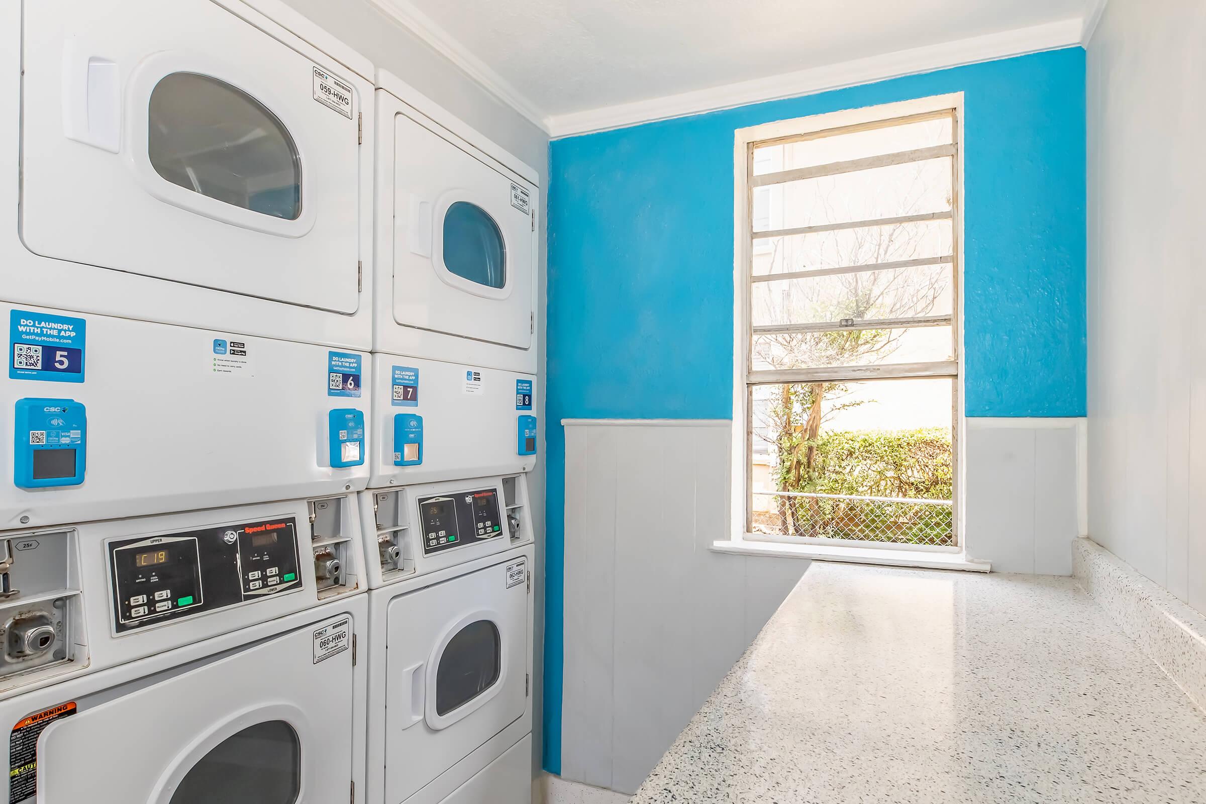 A bright laundry room featuring stacked washing machines and dryers against a blue wall. A window provides natural light, with a view of greenery outside. A countertop is situated beneath the window, offering space for folding laundry. The area appears clean and well-maintained.