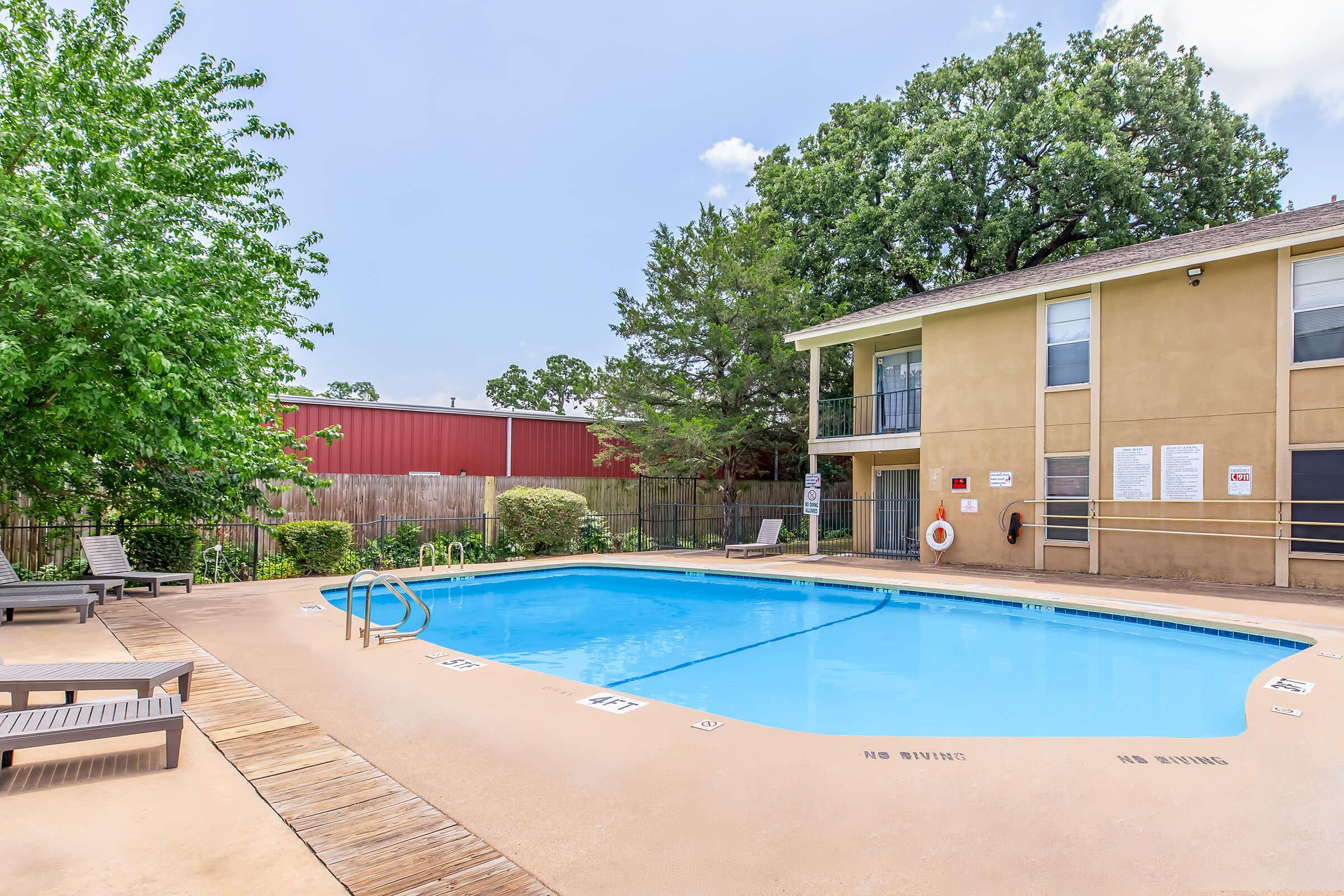A clear swimming pool surrounded by wooden deck chairs, located in a residential area. Lush green trees and a fence provide privacy, while a building with multiple balconies can be seen in the background. The sky is bright with a few clouds, creating a welcoming atmosphere for relaxation and leisure.