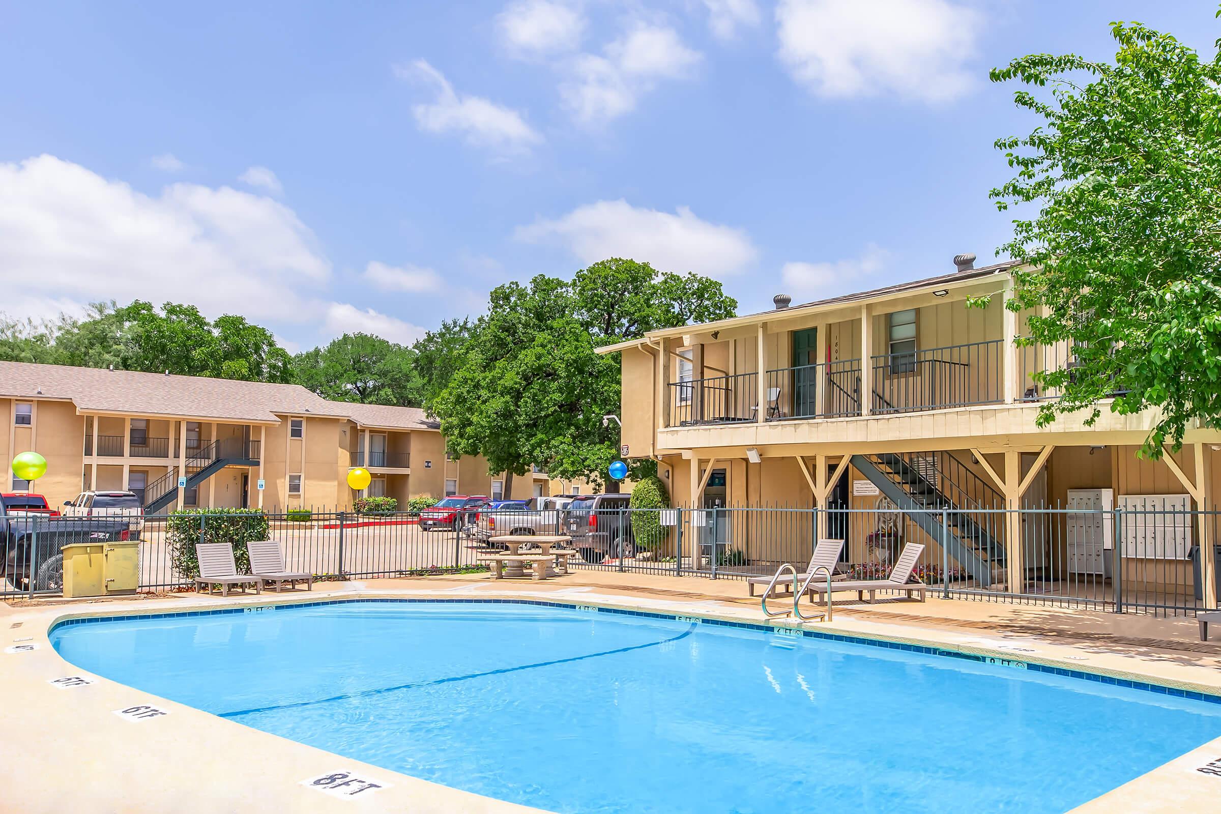A sunny residential area featuring a swimming pool surrounded by lounge chairs. In the background, two-story apartment buildings are visible, along with green trees and a clear blue sky. Yellow balloons add a festive touch to the atmosphere.
