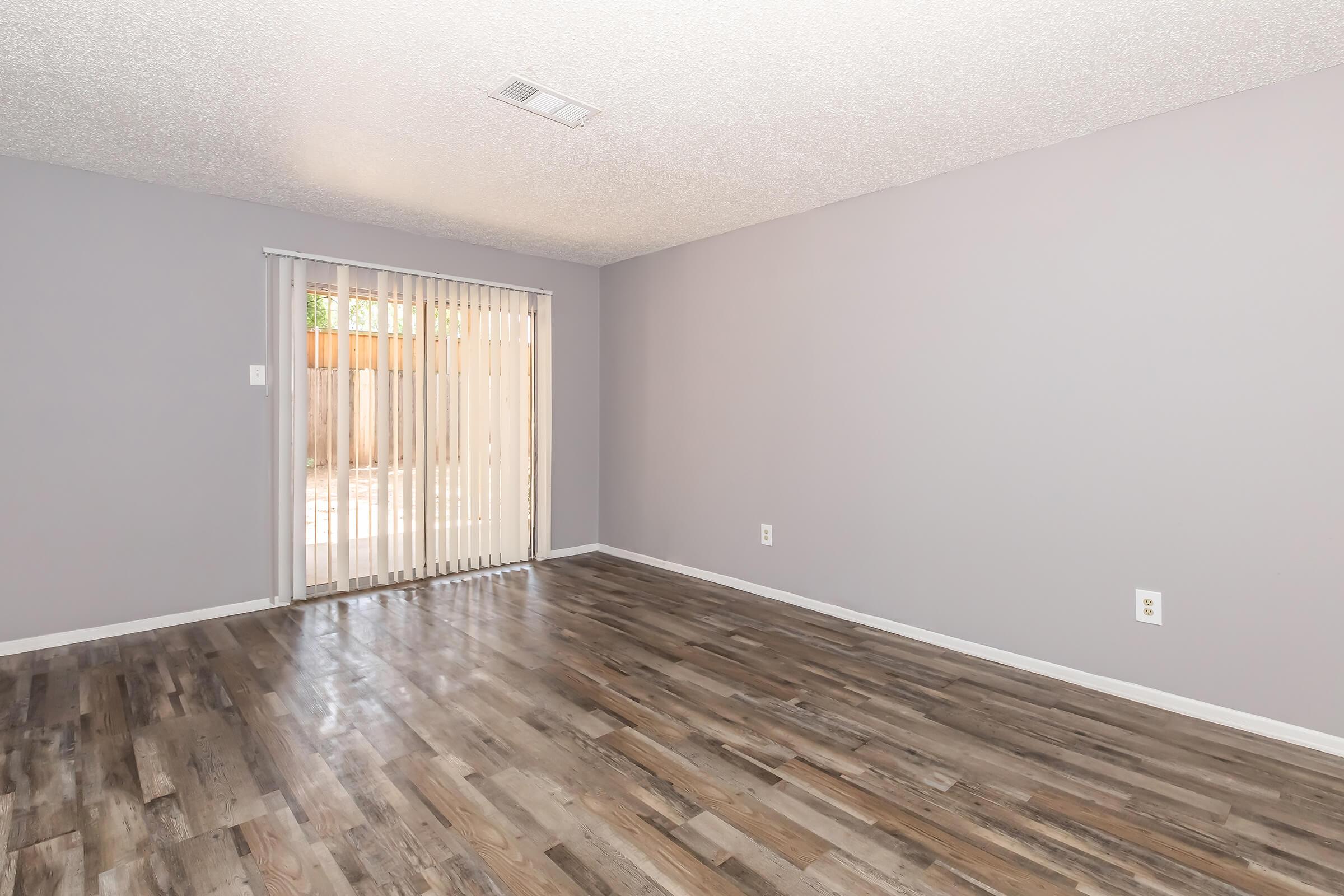 Empty room with light gray walls and hardwood floors. A sliding glass door with vertical blinds lets in natural light, leading to an outdoor space. The room is spacious, featuring clean lines and a minimalist design.