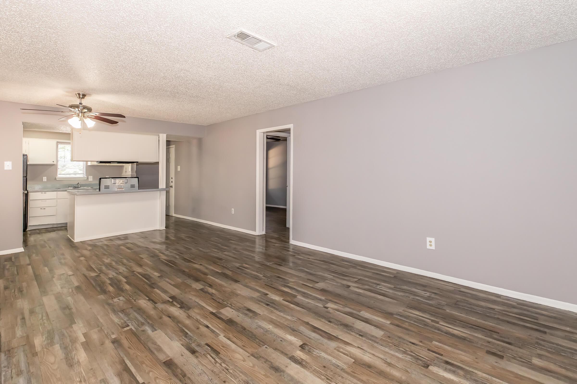 A spacious, light-filled living area featuring a textured ceiling, modern ceiling fan, and laminate flooring. The open layout leads to a kitchen area with white cabinetry, while the walls are painted in soft gray. A doorway on the right leads to another room, adding to the inviting atmosphere.