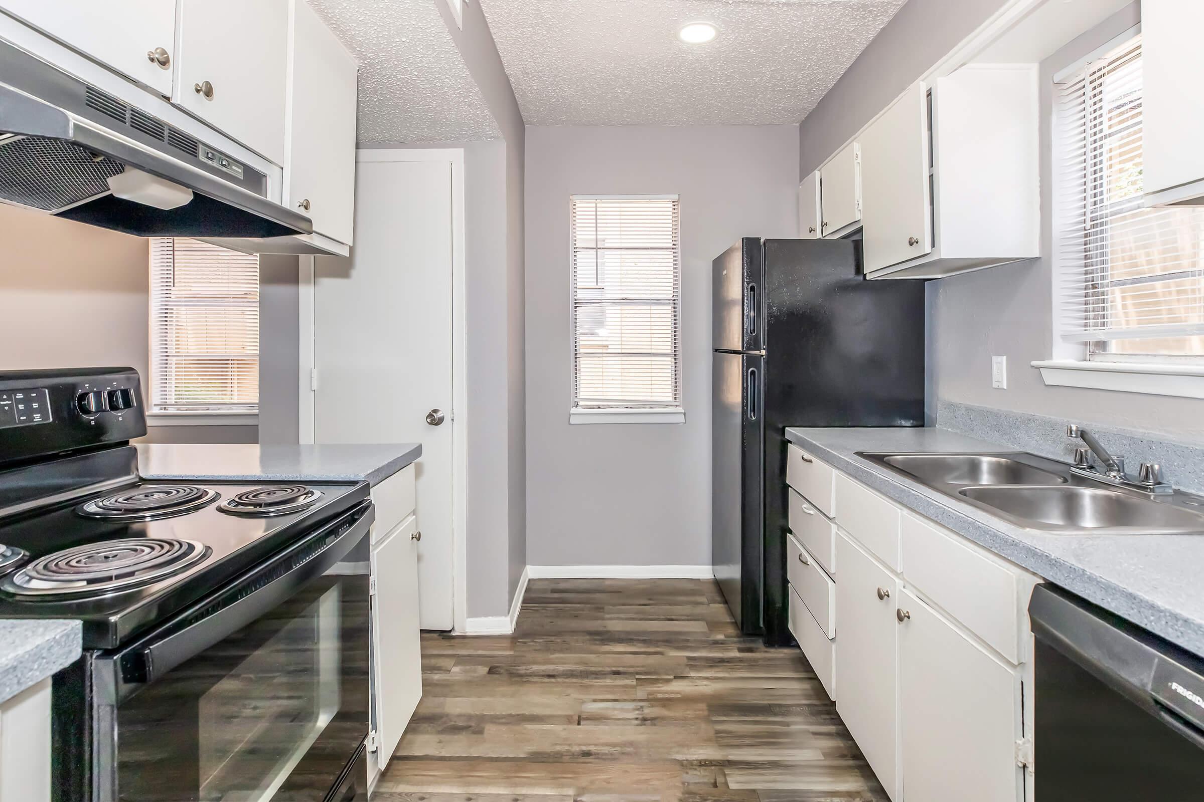 A modern kitchen featuring white cabinetry, a black refrigerator, and an oven with a stovetop. Natural light enters through two windows, highlighting the gray walls and wooden flooring. The layout includes a double sink and ample counter space, creating an organized and inviting cooking area.