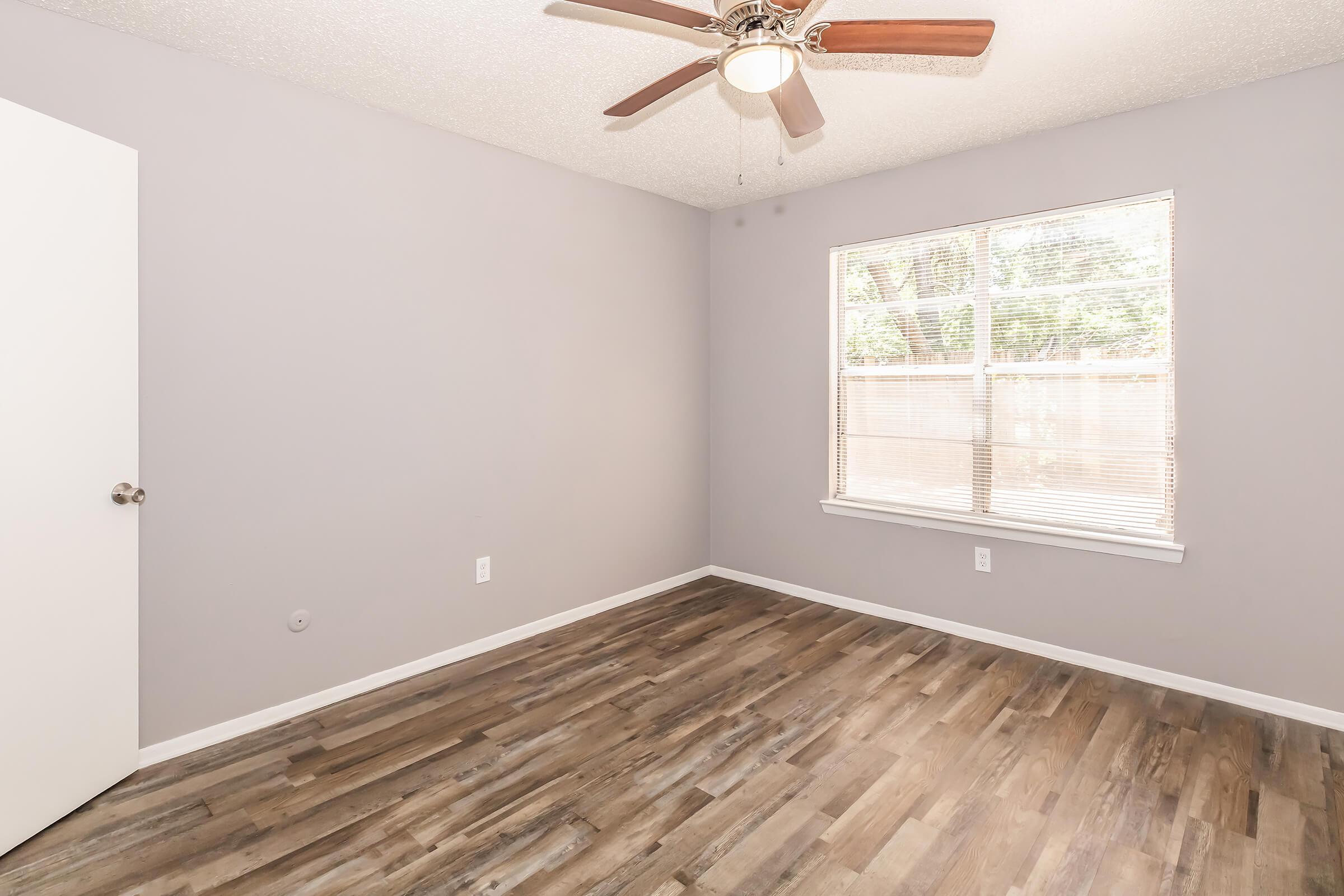 A minimalist bedroom featuring light gray walls, a wooden floor, a ceiling fan with wooden blades, and a window allowing natural light. The room includes a white door and no furniture, creating an open and spacious feel.