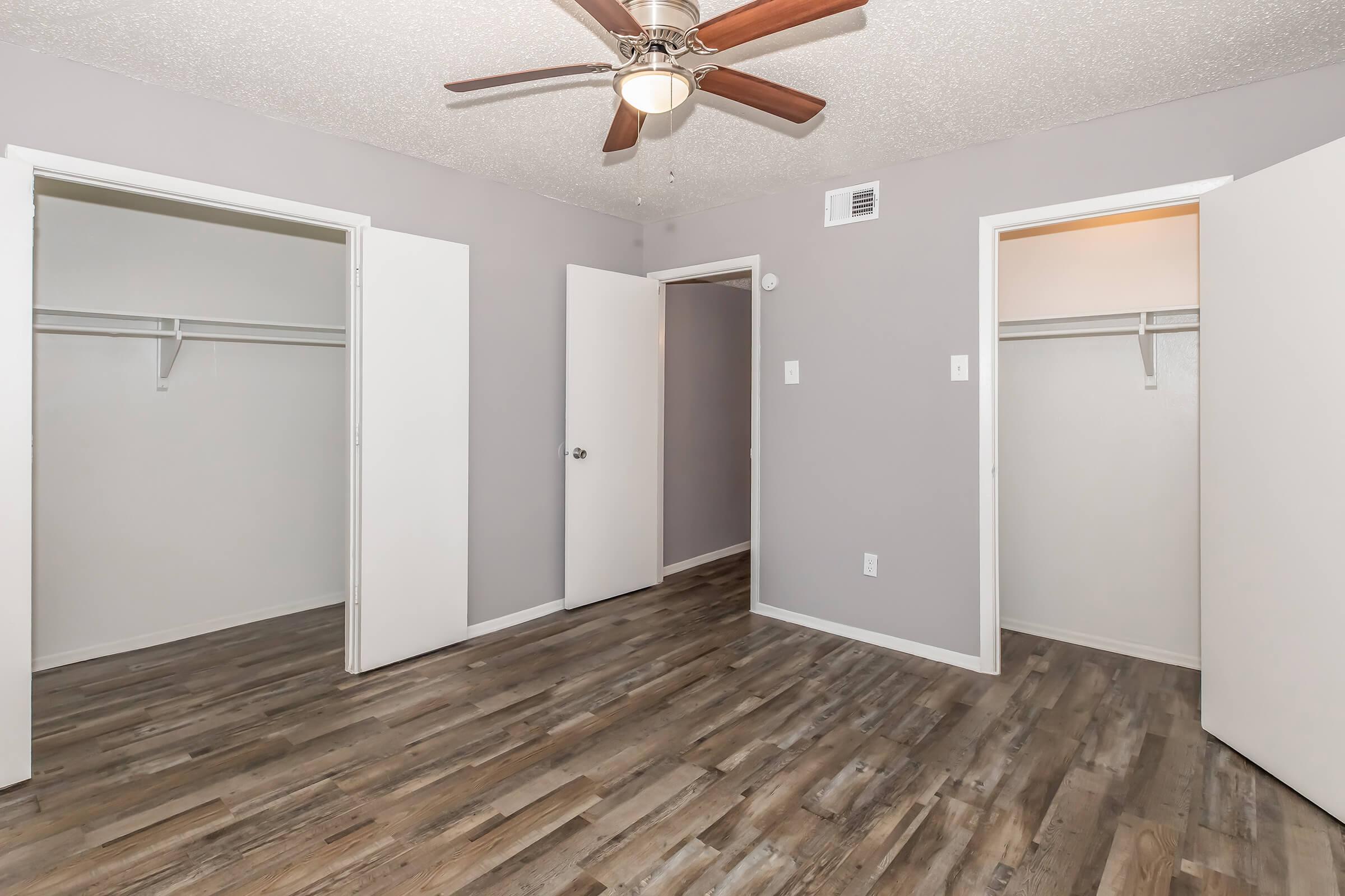 A view of a room featuring a light gray wall color, two open doors leading to closets on either side, and a ceiling fan. The floor is covered with light-colored wood or laminate flooring, providing a spacious and airy feeling.