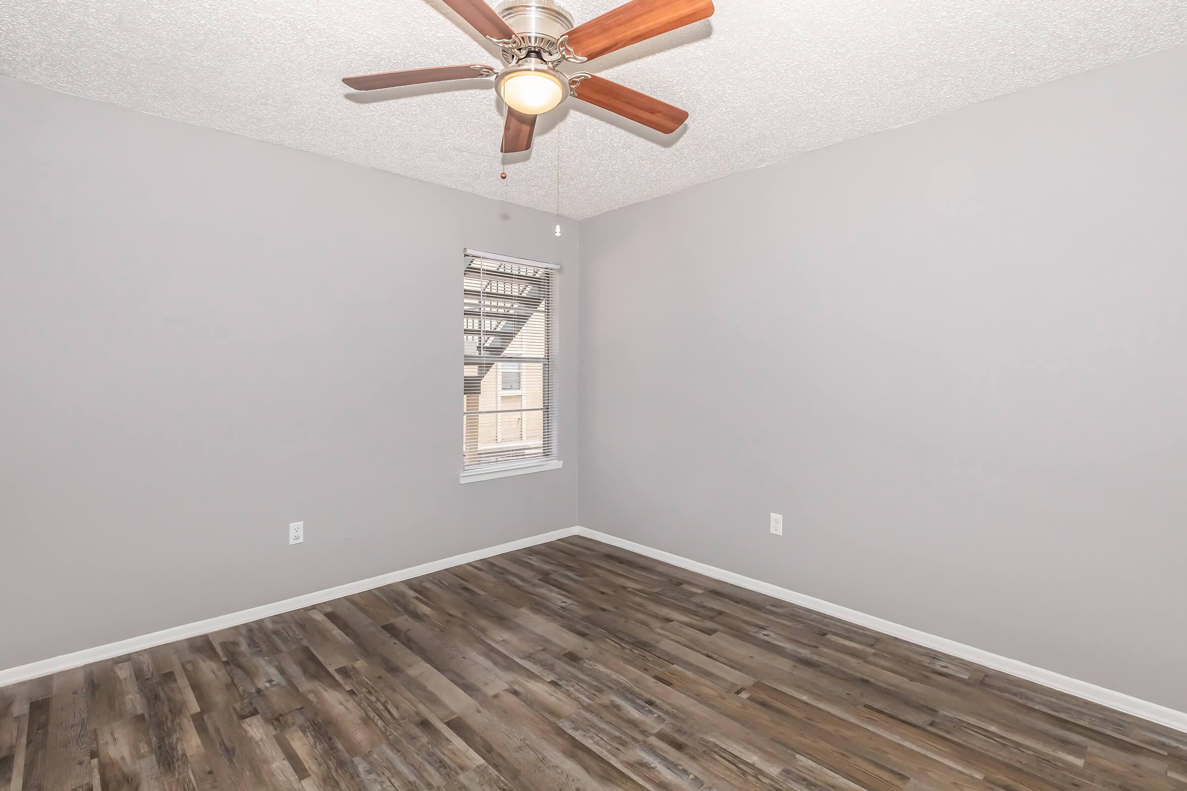A light gray room with hardwood flooring and a ceiling fan. The walls are a soft gray, and there is a window with blinds allowing natural light. The space is empty, making it suitable for various uses or decorating styles.