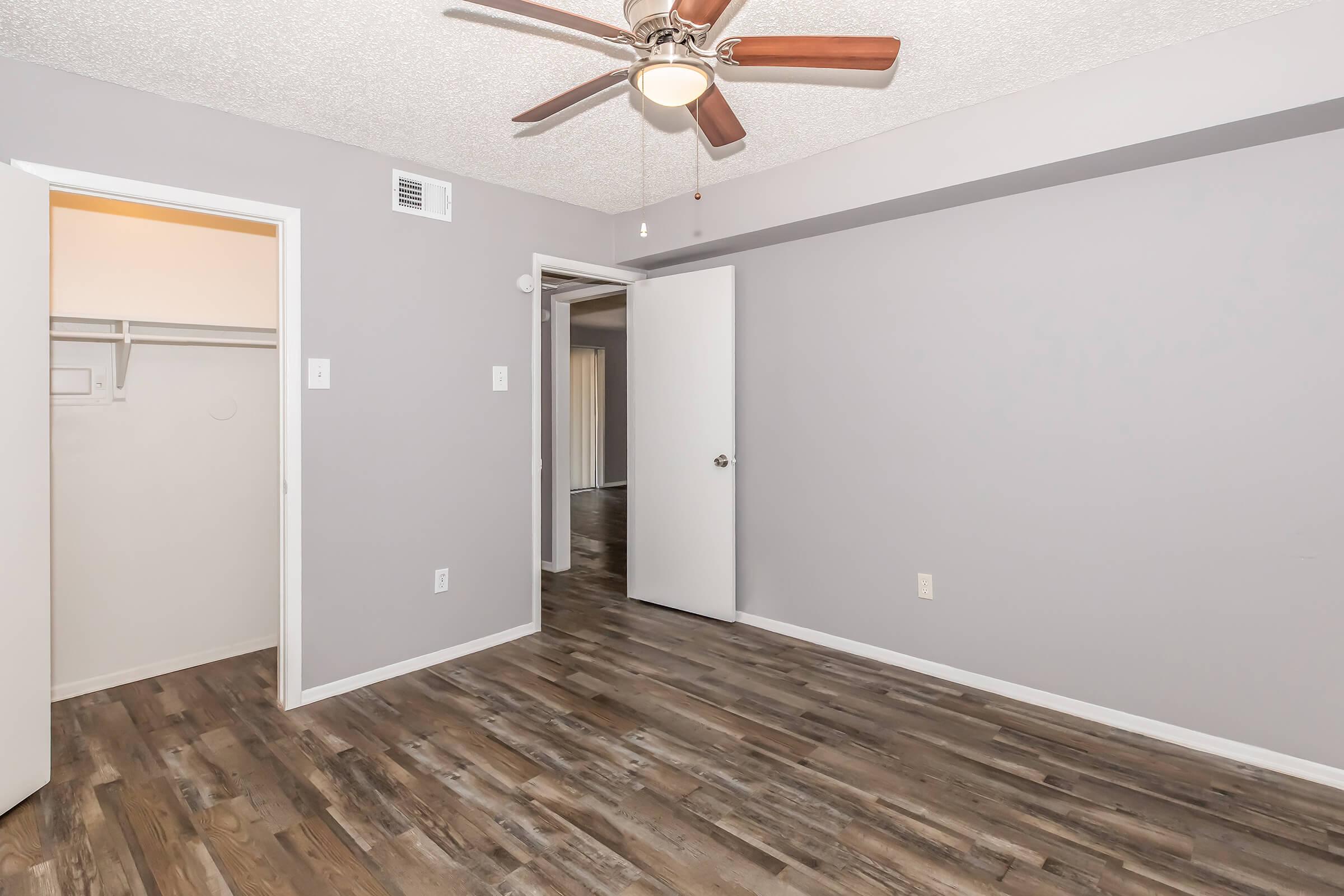 A well-lit room with gray walls and wooden flooring. A ceiling fan is installed, and there are two doors: one leading to a closet and another to an adjacent area. The space appears clean and modern, suitable for various uses.