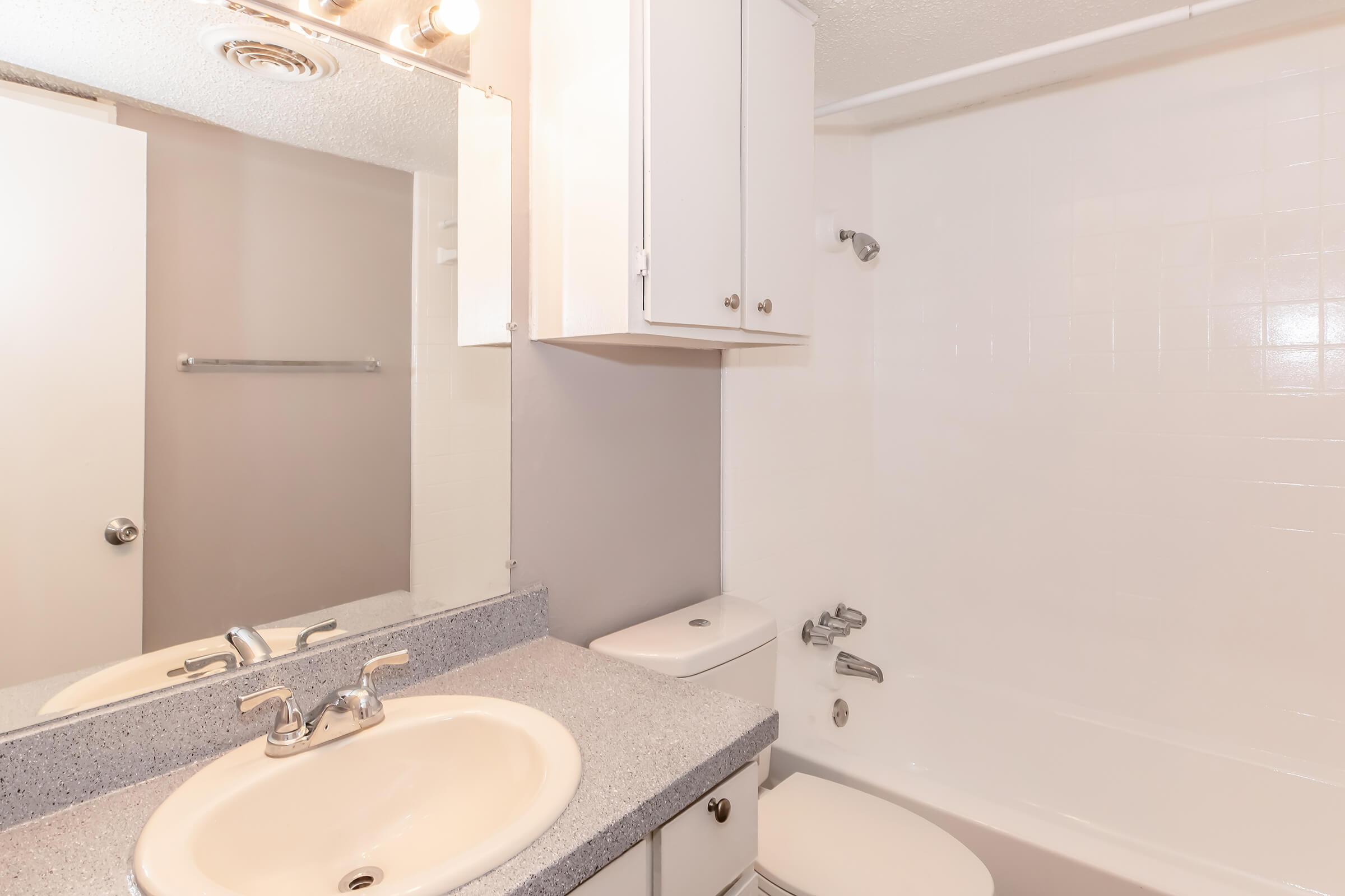 A clean bathroom featuring a white bathtub, a sink with a faucet, and a large mirror above the sink. The walls are a light color, and there are white cabinets for storage. The space is well-lit, creating a bright and inviting atmosphere. A towel rack is also visible on the wall.