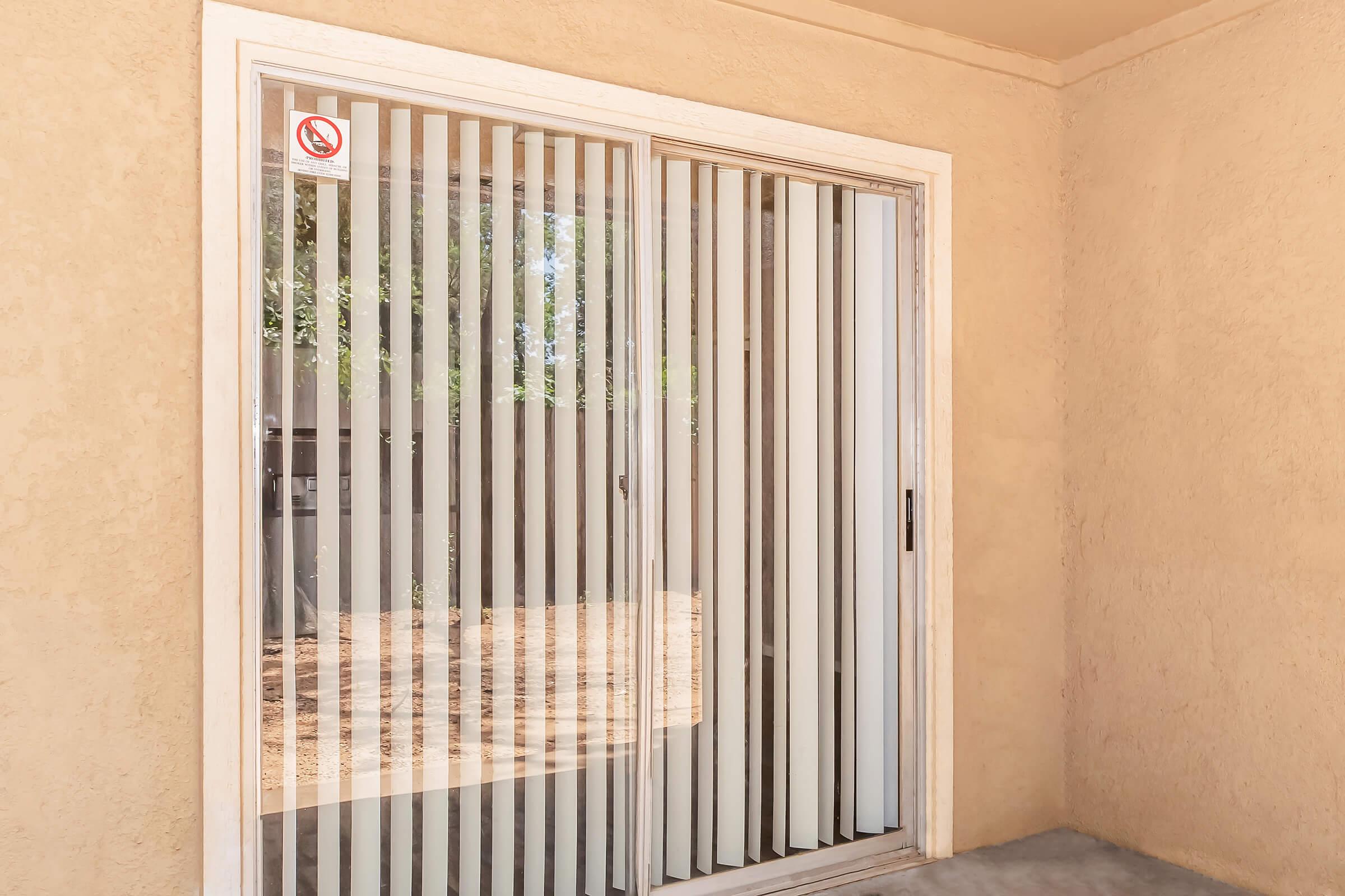 Sliding glass door with vertical blinds, set in a beige wall. The door leads to an outdoor area, with natural light filtering through. There is a no-smoking sign on the glass. Surrounding area appears to be a patio or balcony.