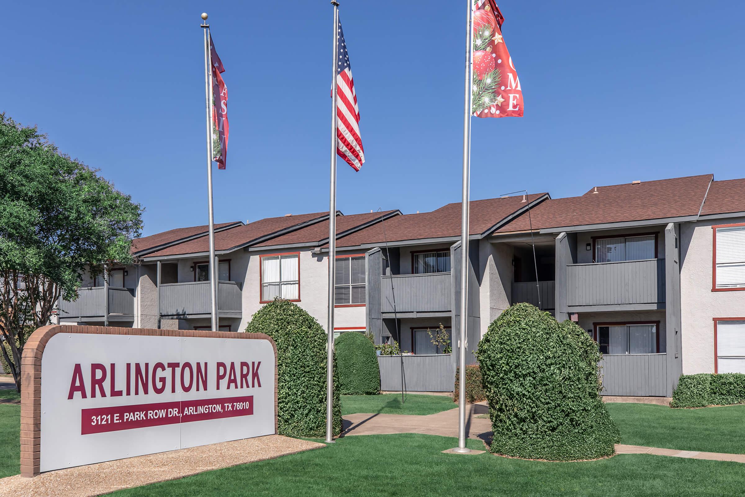 An apartment complex named Arlington Park, featuring a large sign with the name prominently displayed. Below the sign are two flags, and the building has a modern design with multiple windows and balconies. The landscaping includes well-maintained grass and bushes.