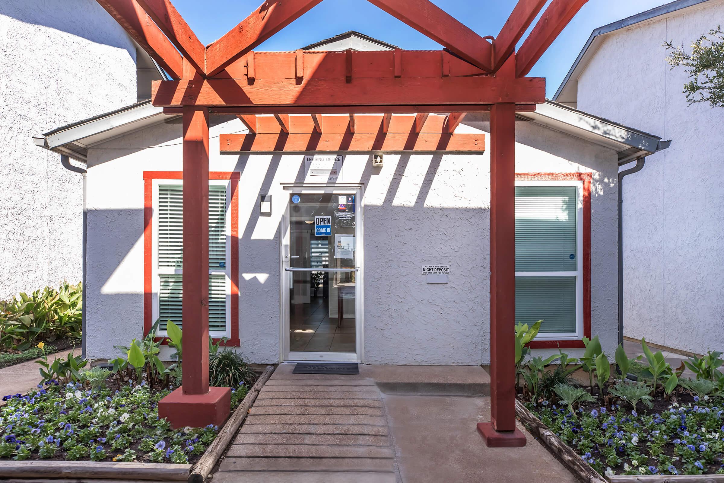 A welcoming entrance to a building, featuring a red wooden pergola over a pathway. The entrance is marked with glass doors and has large windows on either side. Lush green plants and colorful flowers adorn the sides of the pathway, creating a vibrant and inviting atmosphere.