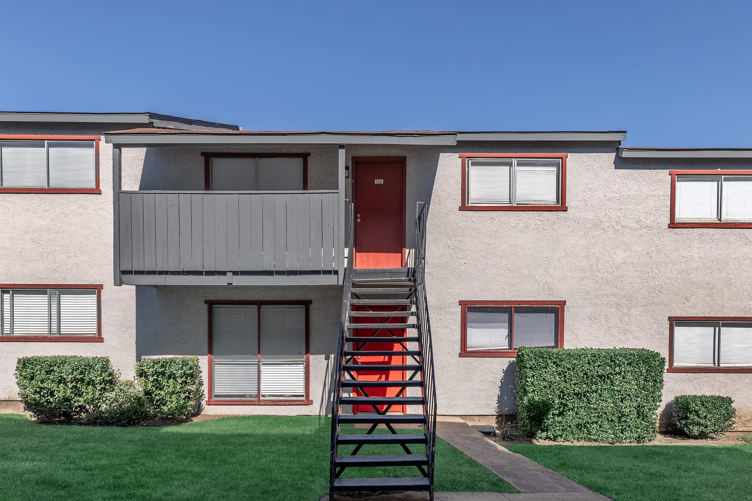 A multistory apartment building with a staircase leading to the second floor. The façade is light-colored with red trim around windows and doors. There is a grassy area in front, with some neatly trimmed bushes on either side of the stairs. The sky is clear and blue.